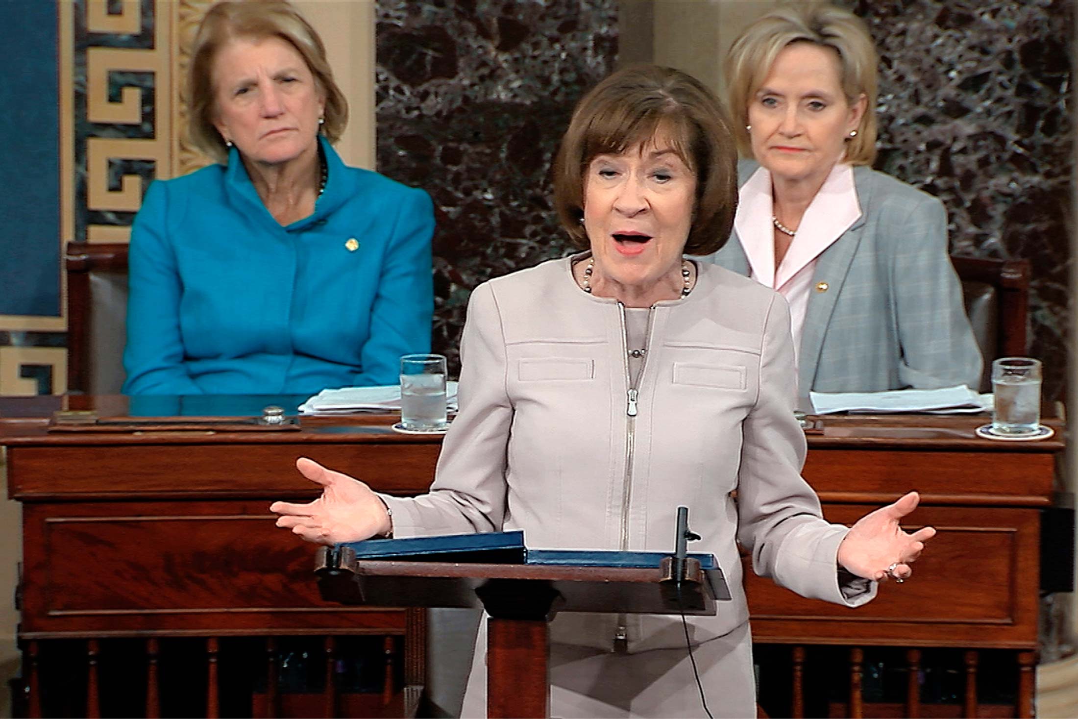 Susan Collins at a podium on the Senate floor with female senators behind her