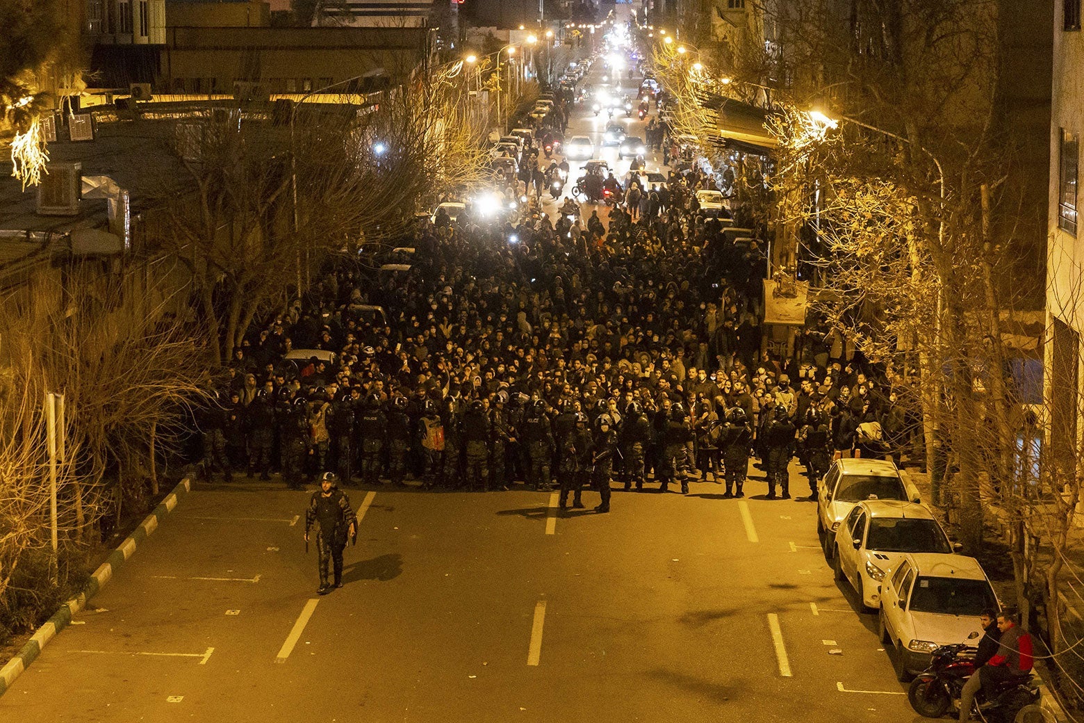 A massive is crowd seen walking on a wide street, led by a cop in riot gear out front. Behind the protesters, cars stand, shining their headlights.
