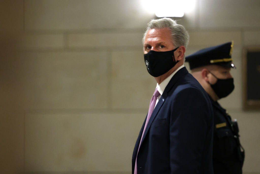 McCarthy wearing a black mask, in profile in a hallway with a masked police officer behind him