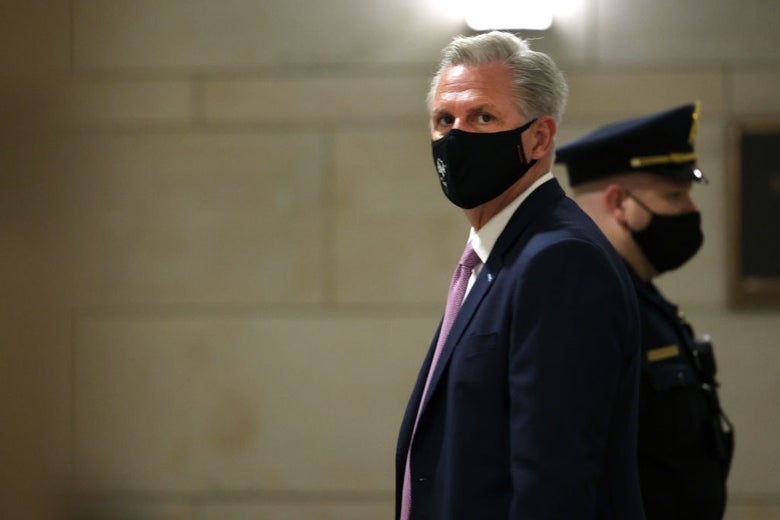 McCarthy wearing a black mask, in profile in a hallway with a masked police officer behind him