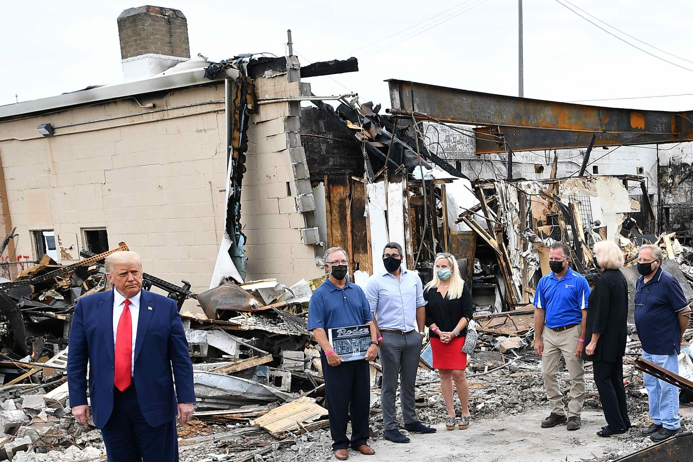 Trump stands before a wrecked building, next to people in masks.