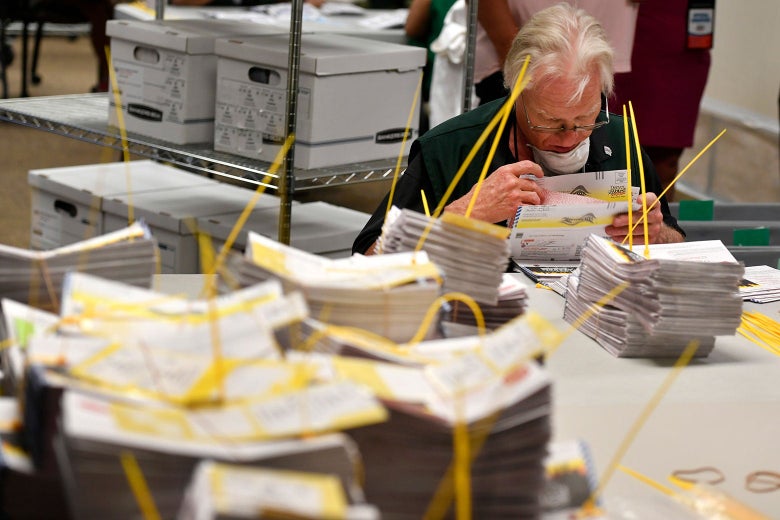 Man sifting through a stack of ballots, sitting at a table covered in zip tied stacks of ballots