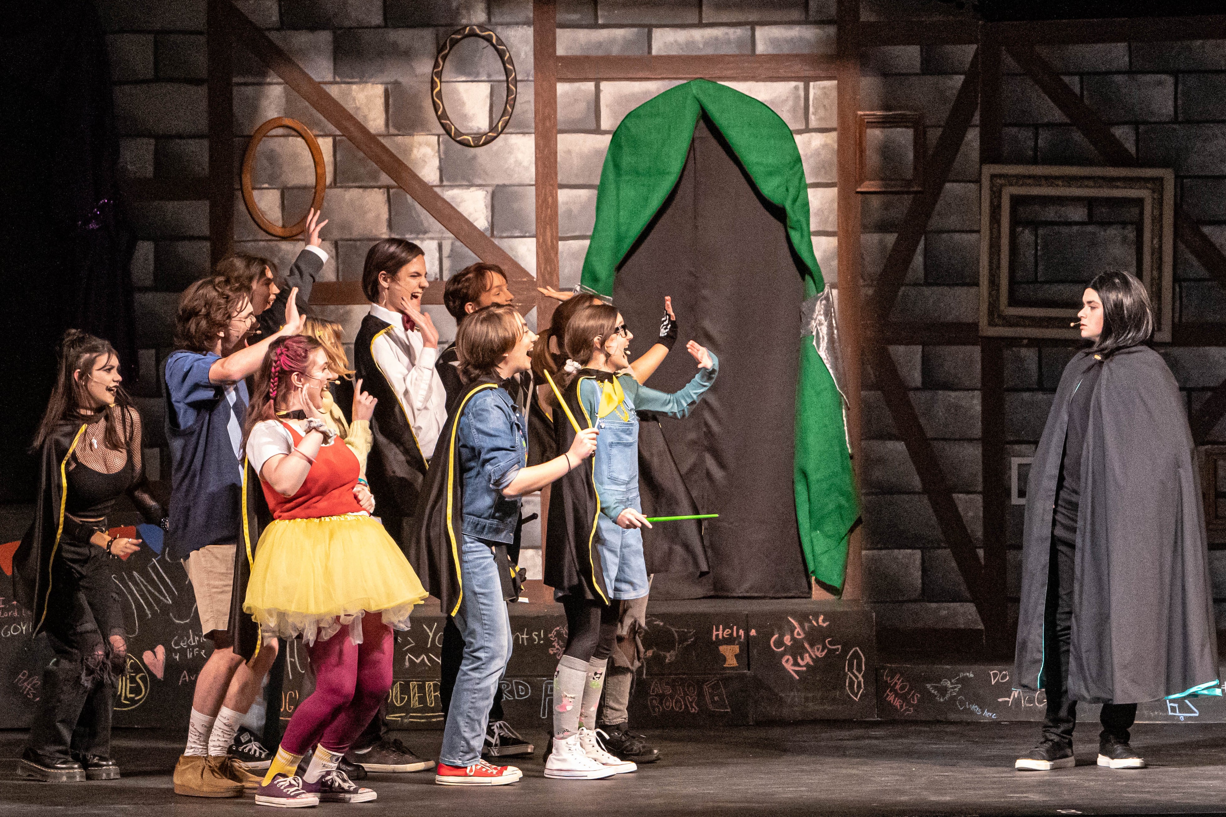 On a stage, a number of kids dressed in silly fashion raise their hands in Potions class.