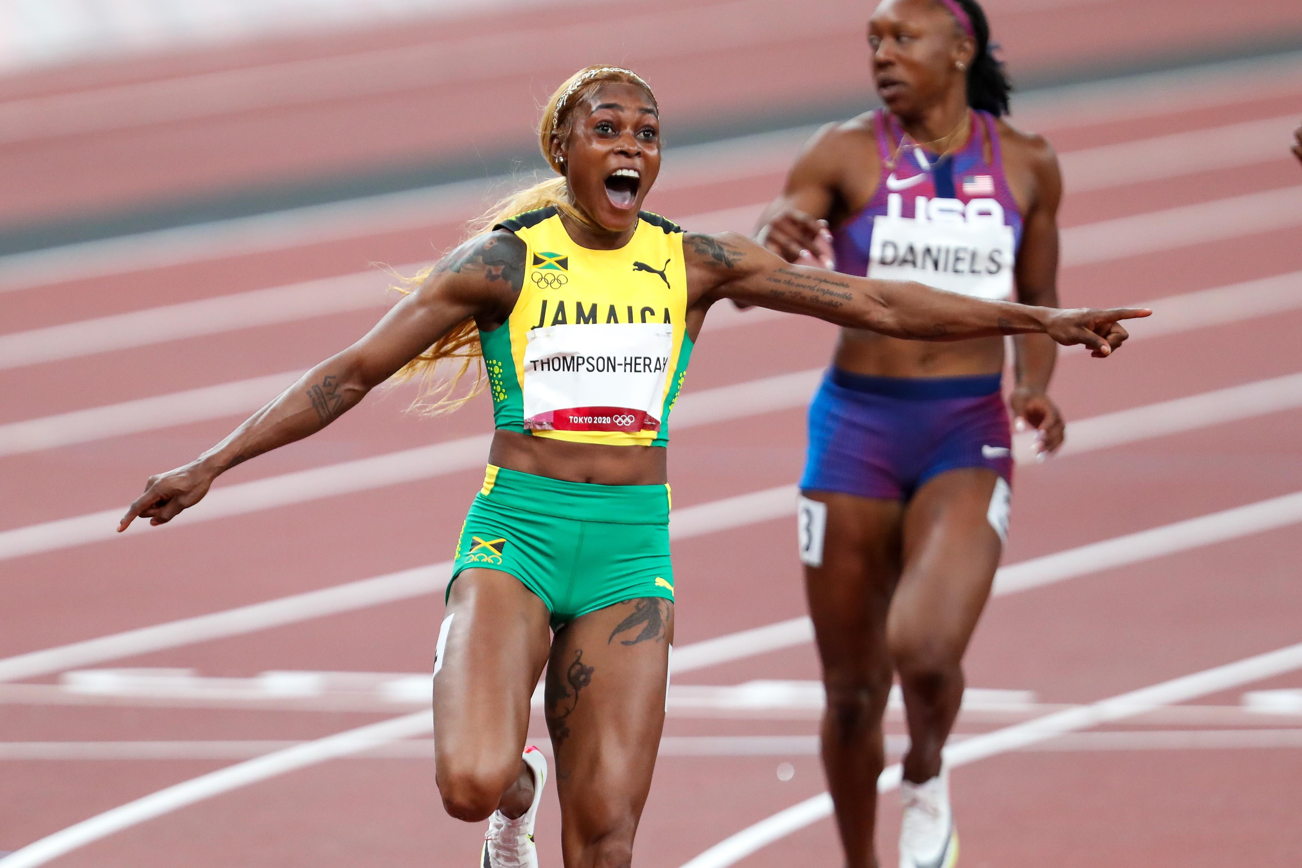 Thompson-Herah yells in celebration with her arms out to the side on the track, American Teahna Daniels in the background