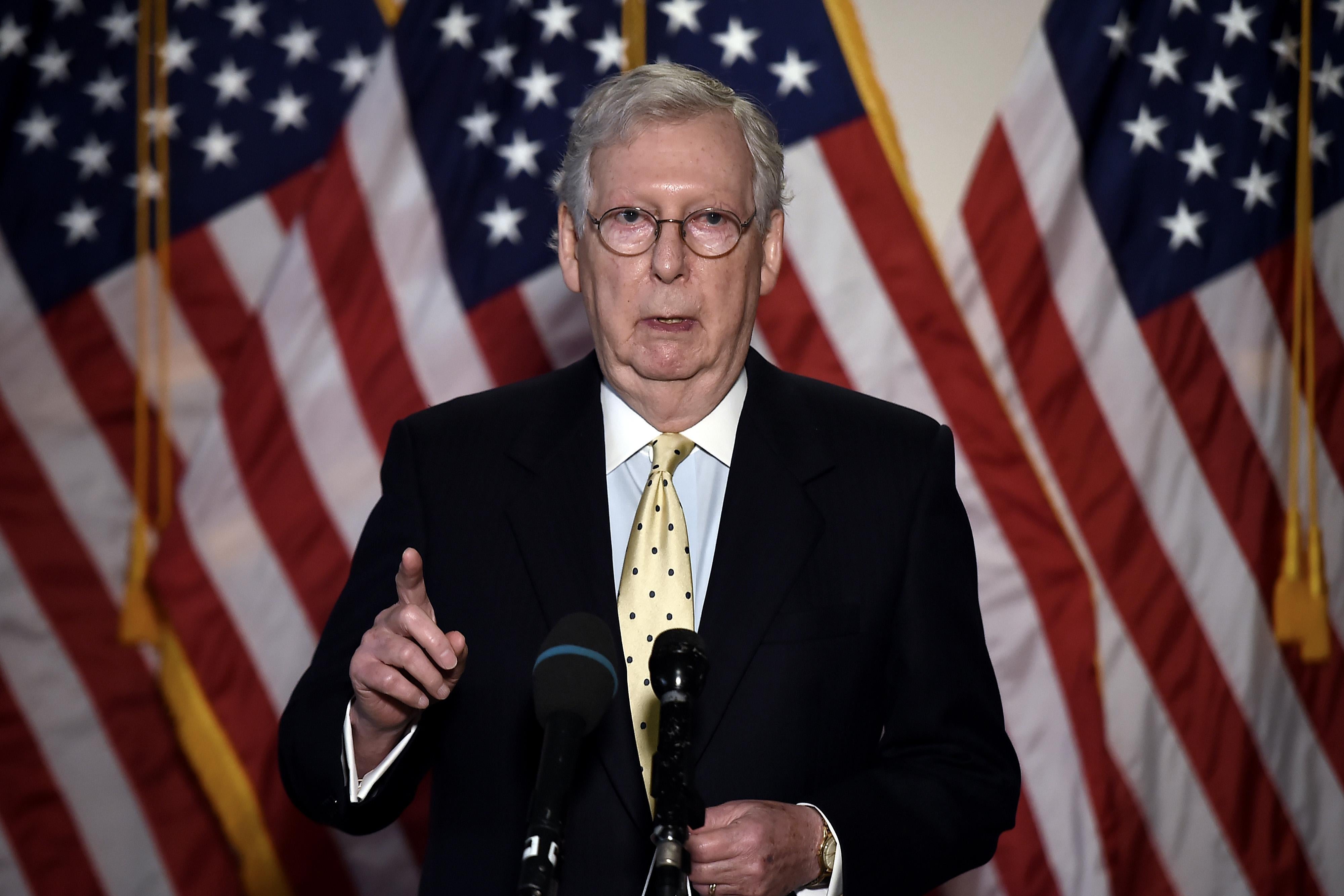 Mitch McConnell speaks into a microphone and points with American flags behind him.