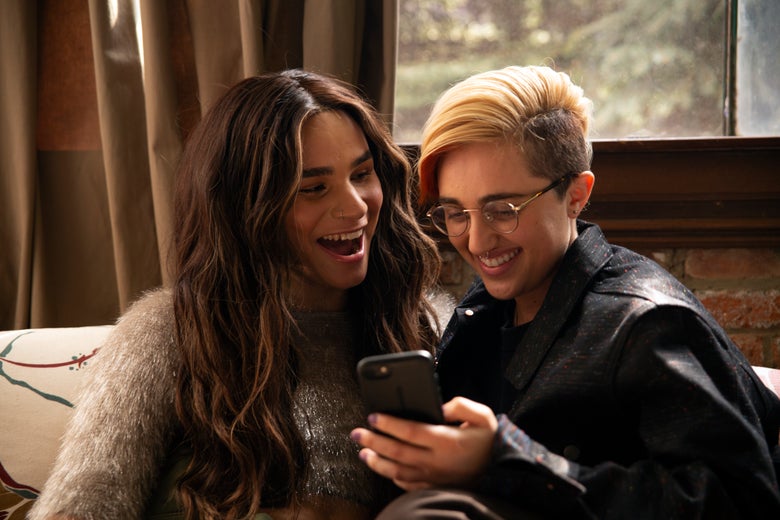A transfeminine non-binary person and transmasculine gender-nonconforming person looking at a phone and laughing.