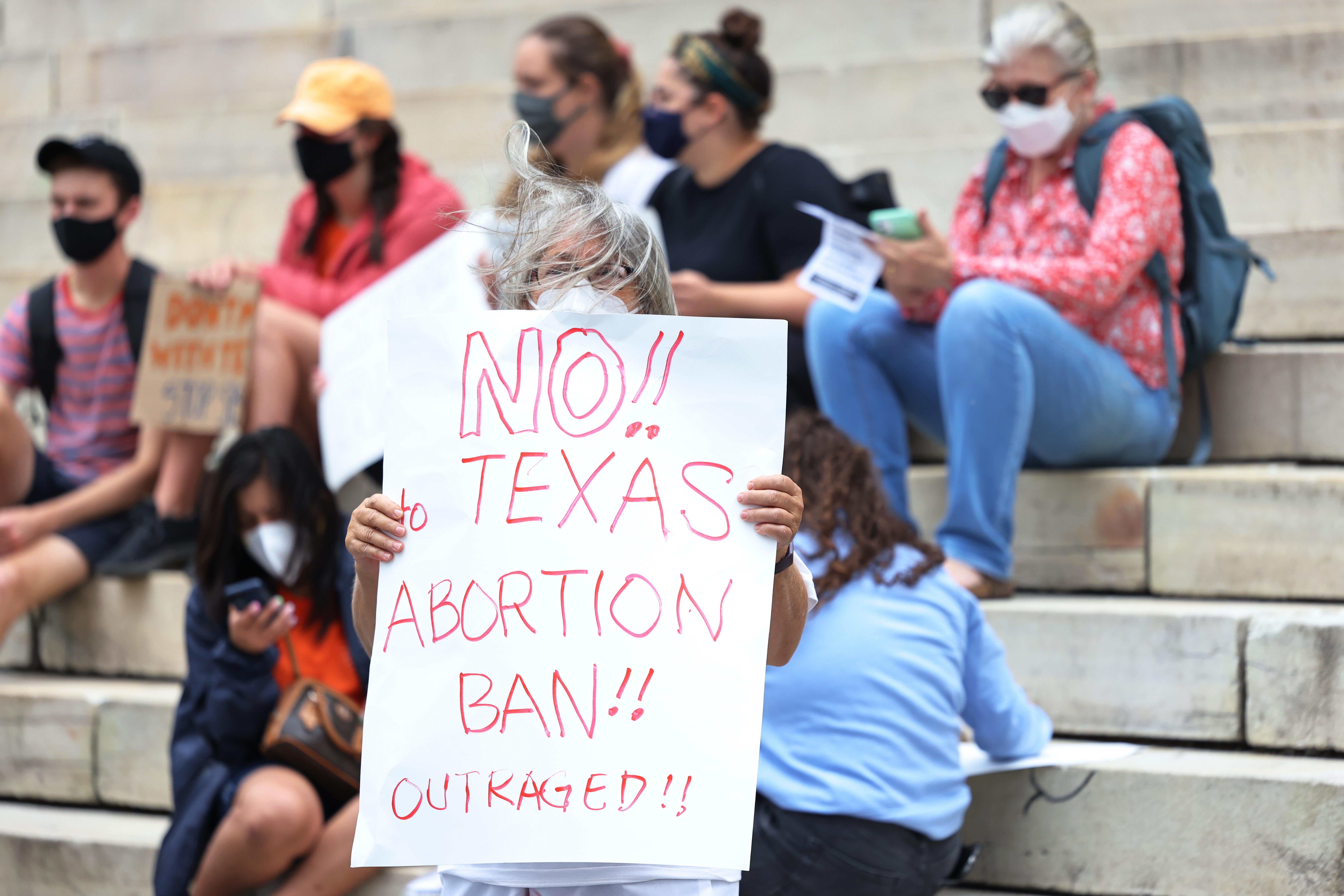 People in masks sitting on steps, with one woman holding a sign reading, "No!! Texas Abortion Ban!! Outraged!!"