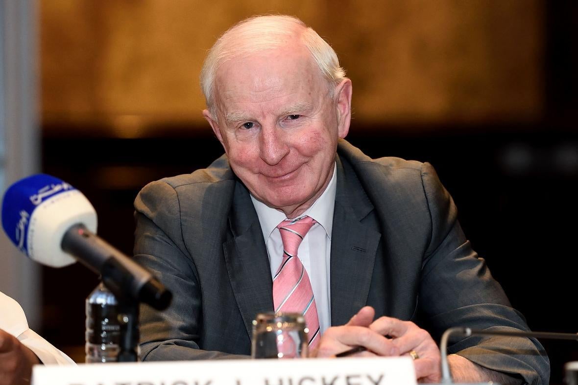Hickey sitting with his hands steepled at a dais and smiling