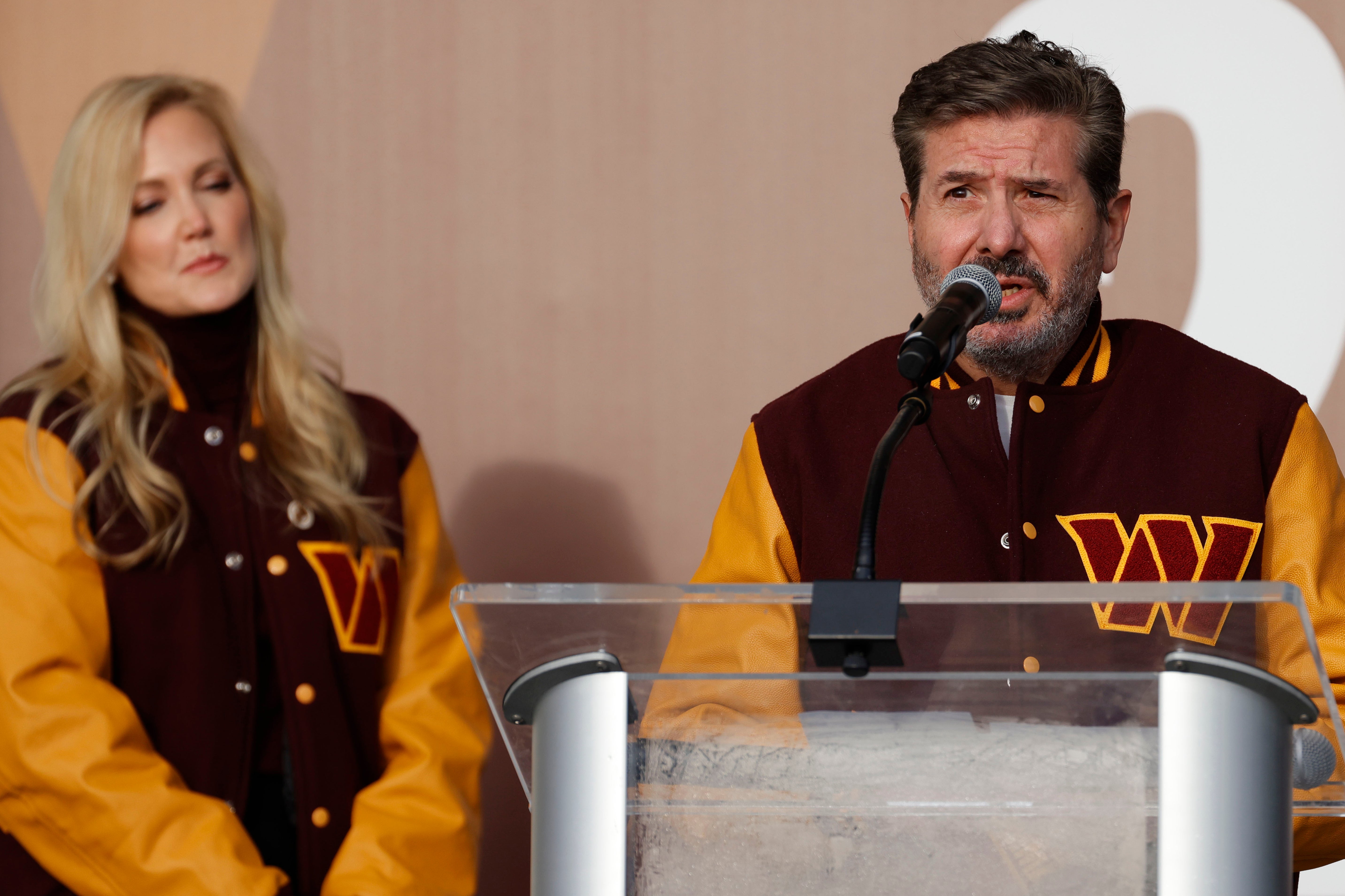 Dan Snyder, in a Washington Commanders jacket, speaks at a podium as his wife Tanya Snyder stands behind him in a matching jacket