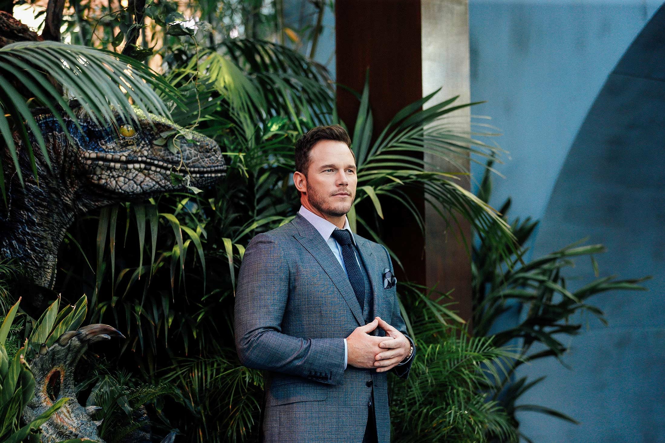 Chris Pratt stands looking solemn, wearing a suit with hands touching and thumbs tented, in front of a backdrop of tropical plants.