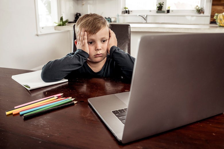 A kid sits at a table with a workbook, colored pencils, and a laptop. He has his hands to his head in frustration.
