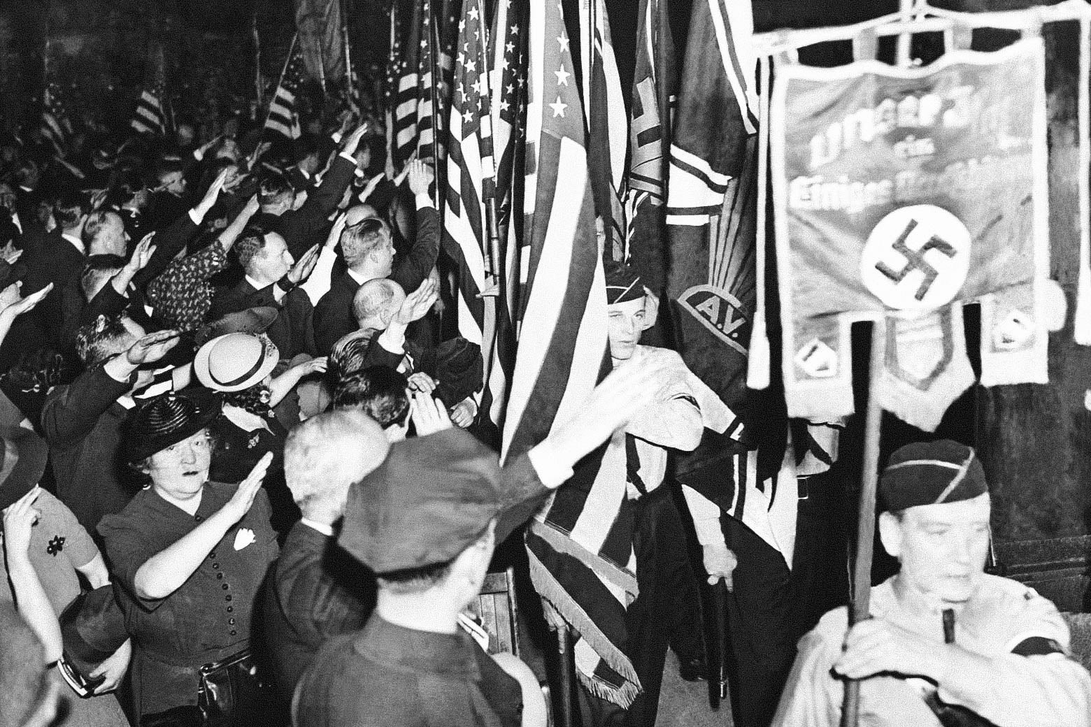 A black-and-white photo shows a crowd giving a Nazi salute to a swastika banner as it is carried past a row of U.S. flags.