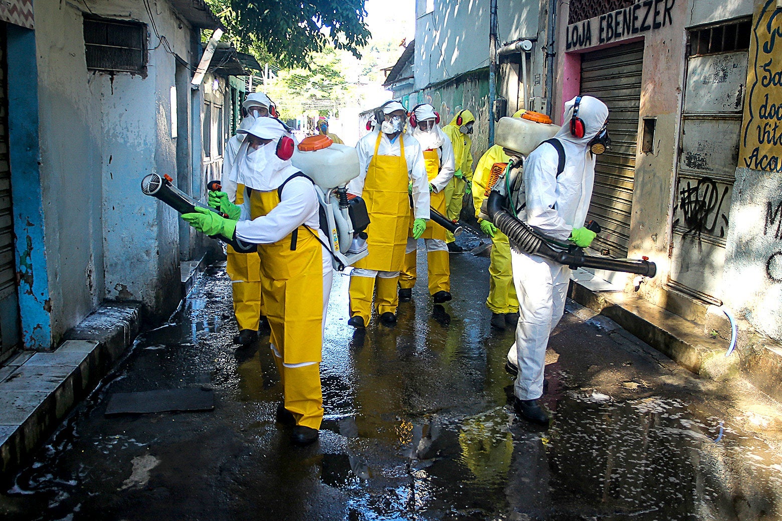 People wearing protective suits and masks and headphones carry vacuums while walking down the aisle of a favela.