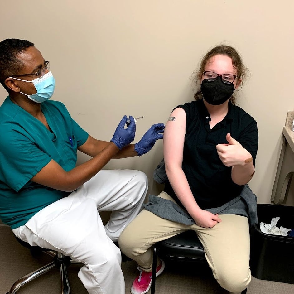 A girl sitting on a chair gives a thumbs-up while a nurse holds a needle.