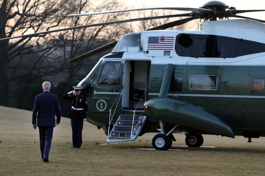 Biden walks toward the Marine One helicopter as a Marine salutes him in gray early morning light.
