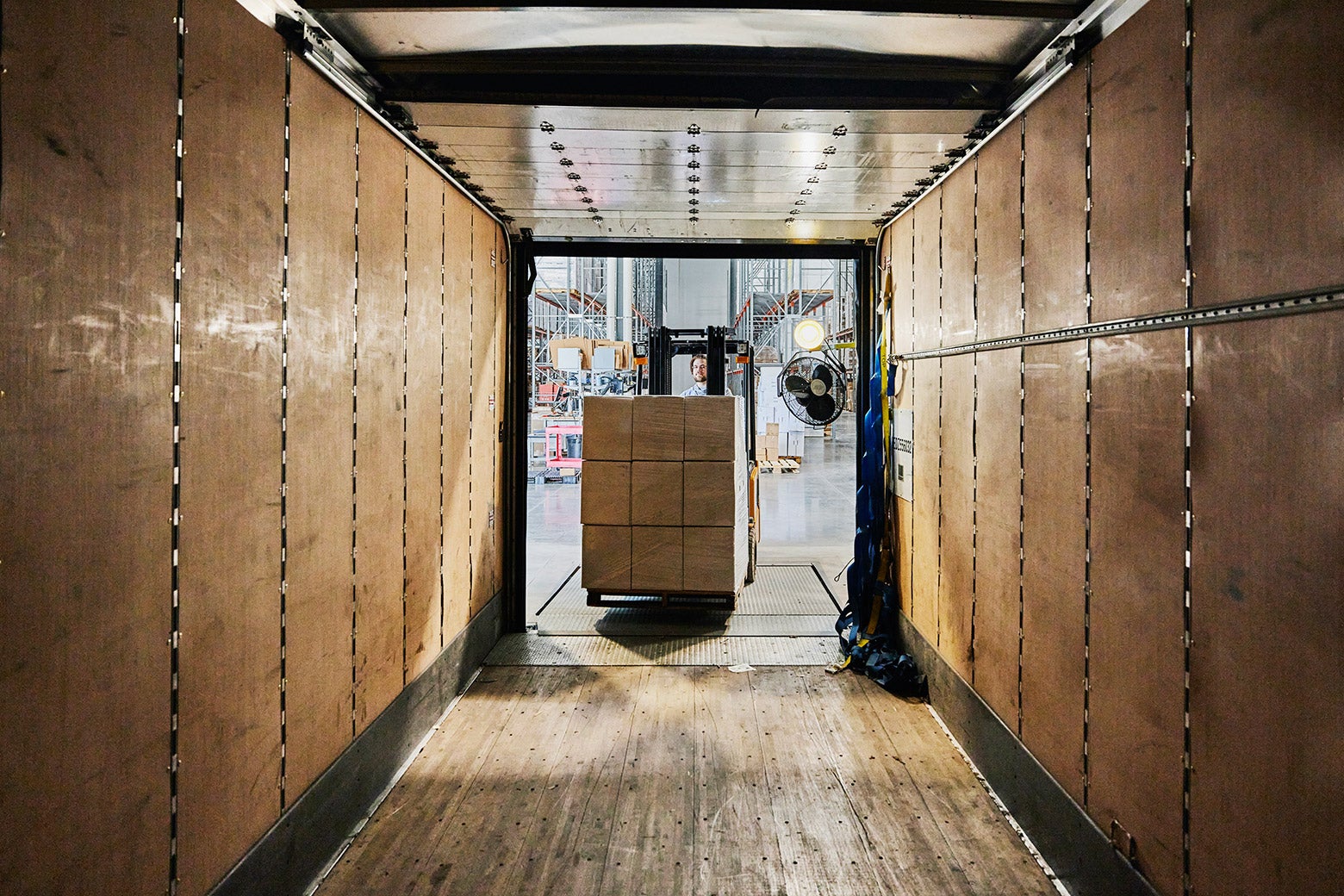 A wide view from inside of truck trailer of a warehouse worker using a forklift to load a pallet of boxes.