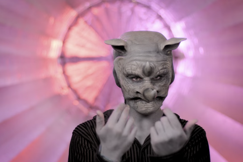 Man in black shirt wearing a rhino mask and pointing to himself.