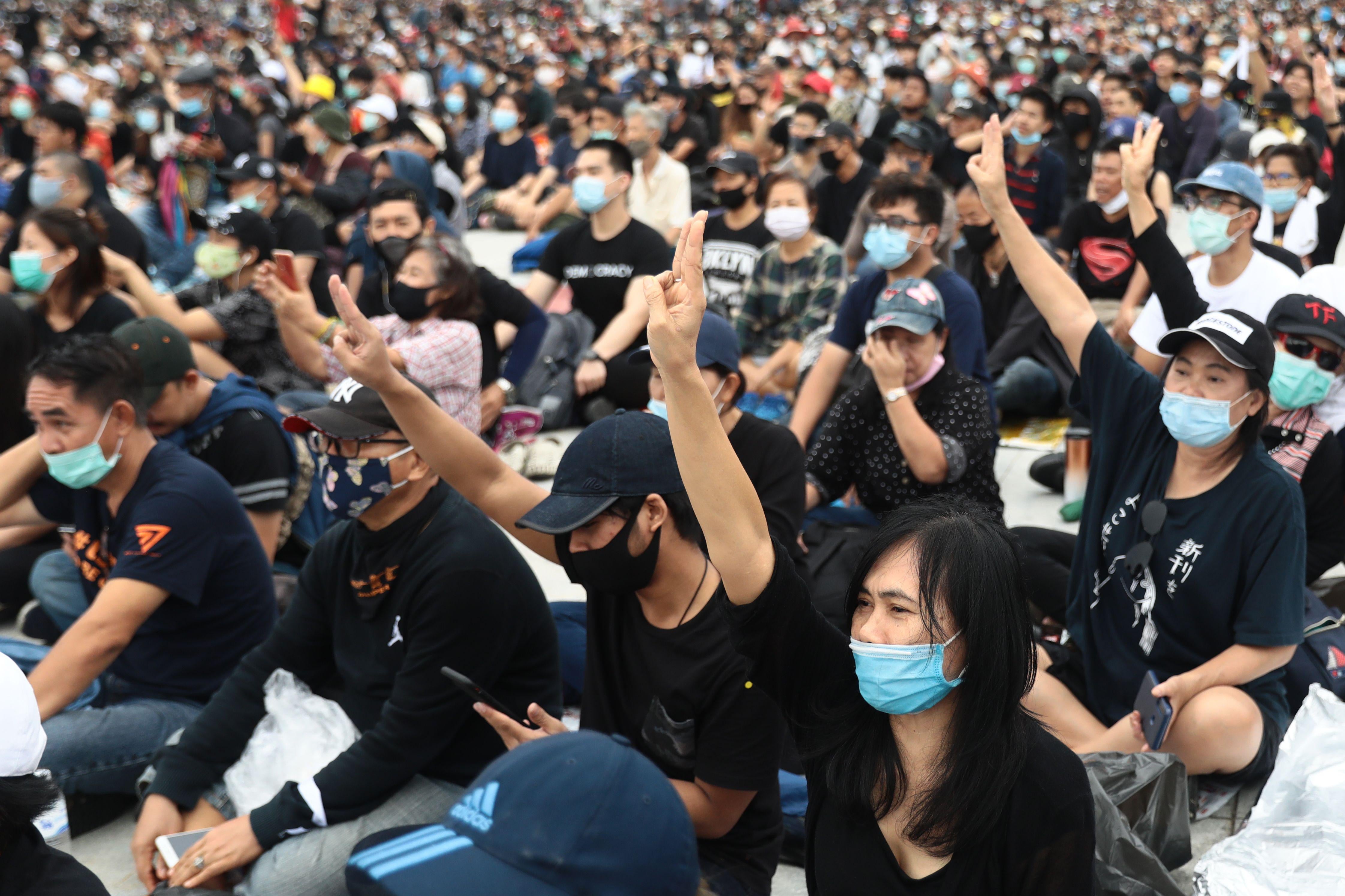 A large crowd of protesters sitting on the ground outside, wearing black T-shirts and masks. Many raise their right arms, holding up three fingers in the Hunger Games salute.