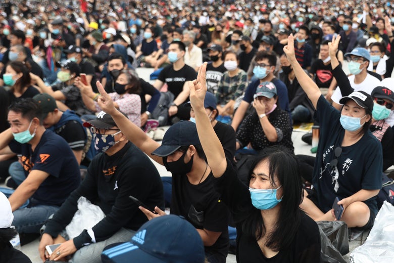 A large crowd of protesters sitting on the ground outside, wearing black T-shirts and masks. Many raise their right arms, holding up three fingers in the Hunger Games salute.