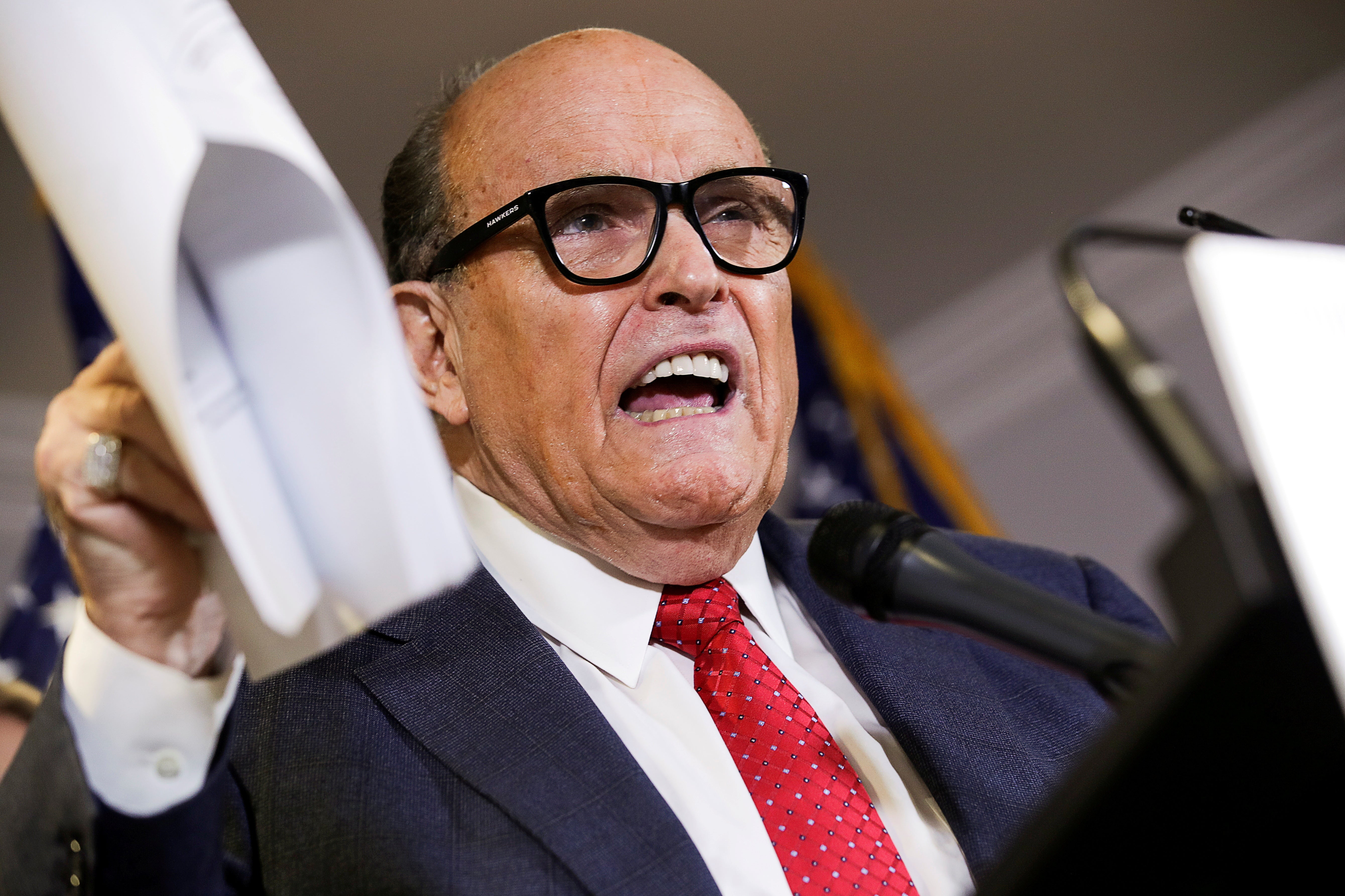 Giuliani speaks at a podium while holding up a packet of papers in one hand