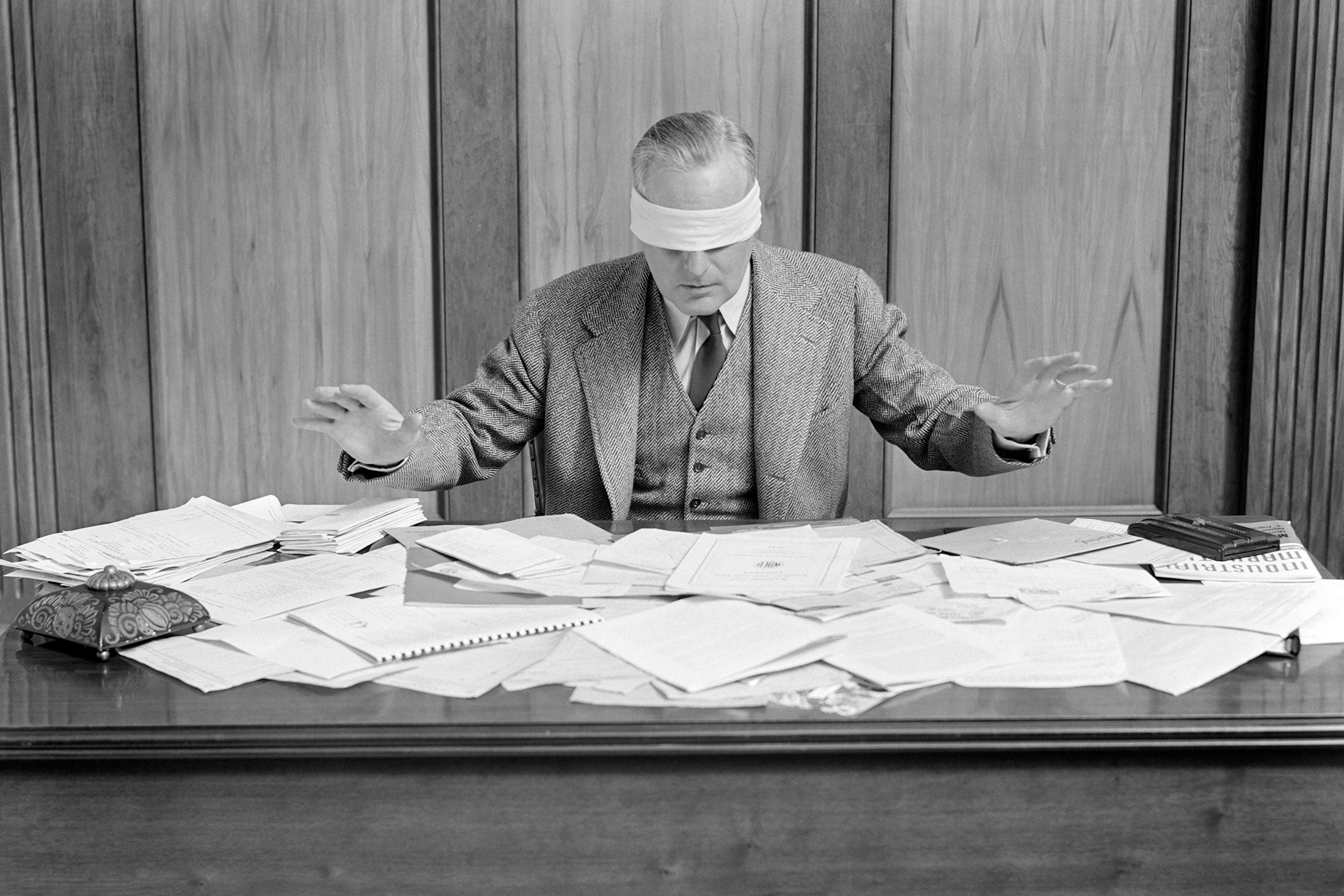 Blindfolded man sitting at a desk covered in papers.