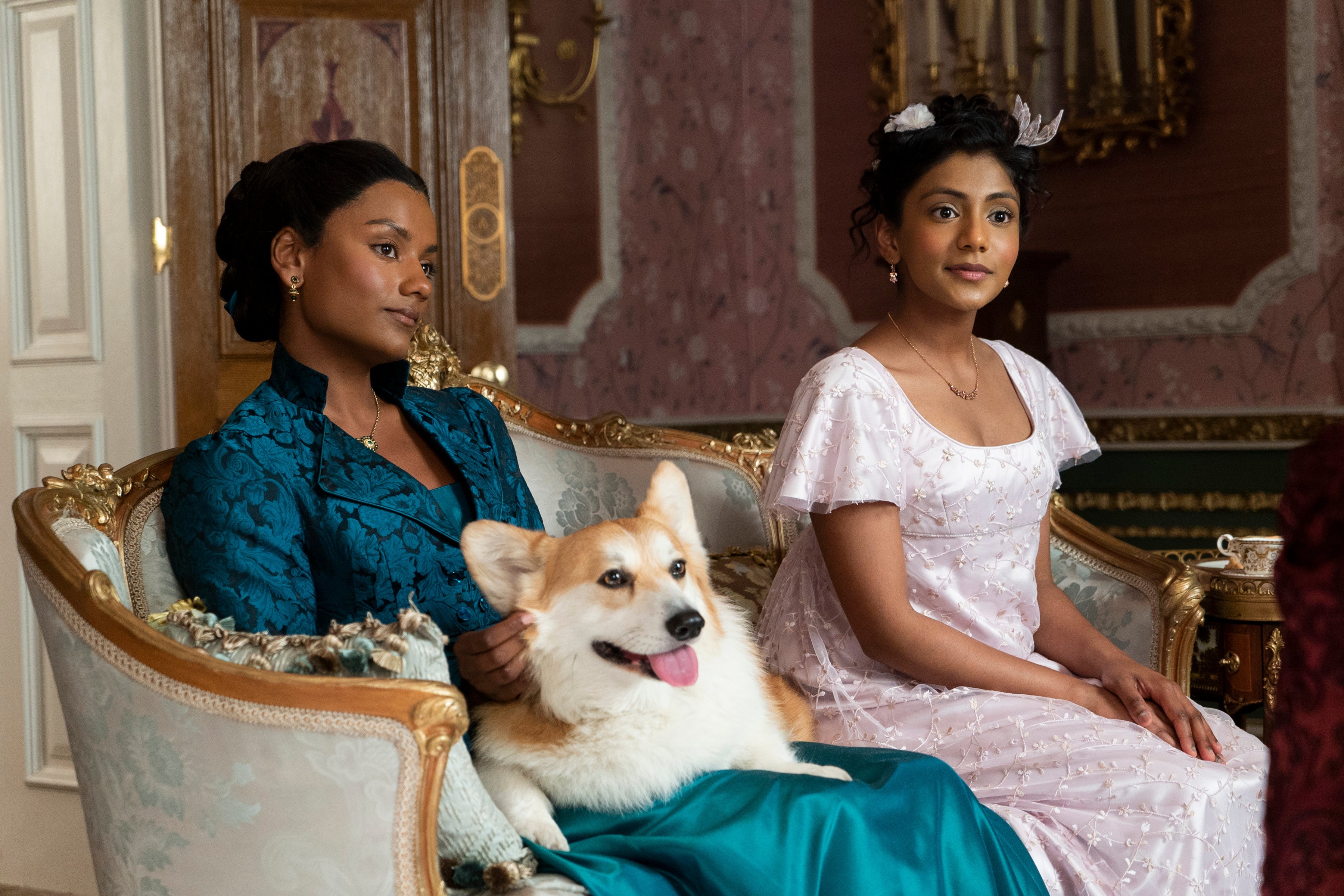 Simone Ashley, with a large corgi in her lap, sits on a couch next to Charithra Chandran.
