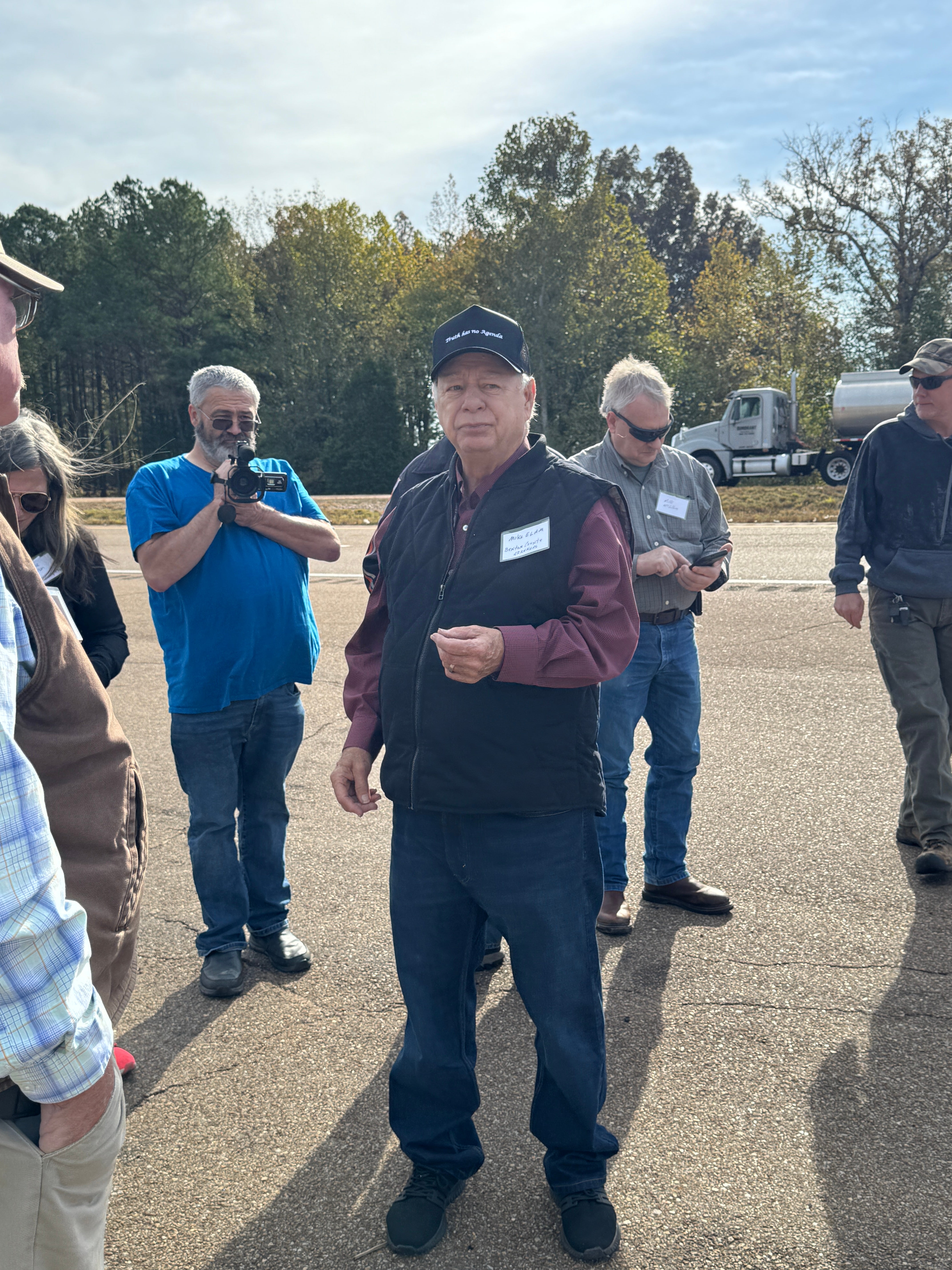 A man in a hat speaking in the center of a group of people in a parking lot.