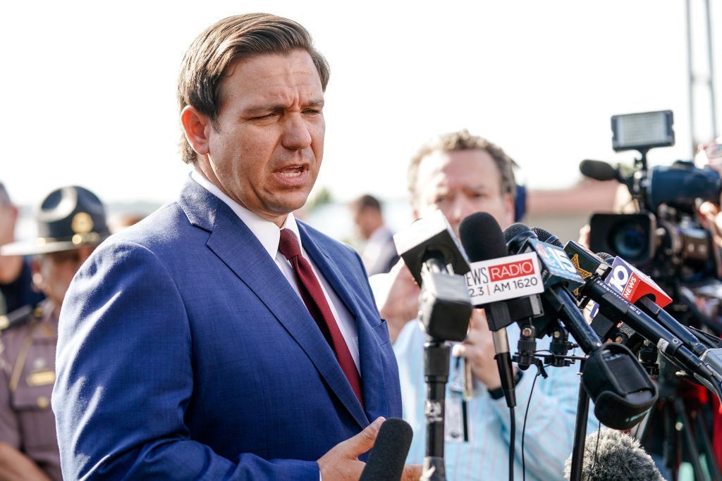 DeSantis, wearing a blue suit and surrounded by members of the media, speaks into a stand of microphones that have been set up outdoors.