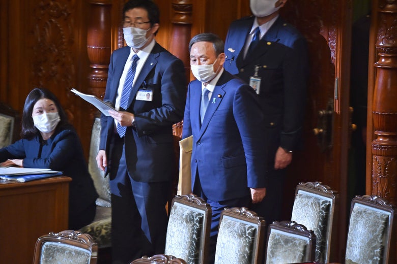 A masked Suga enters parliament surrounded by aides. 