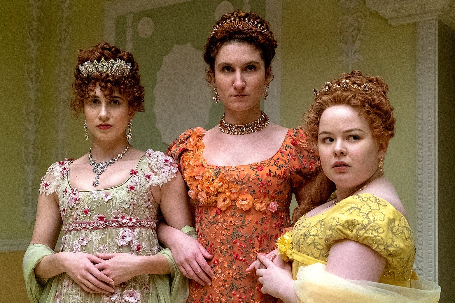 Harriet Cains, Bessie Carter, and Nicola Coughlan wear gowns covered in embroidered flowers and sport oversized, glittering necklaces, earrings, and tiaras. All three have red hair worn up with tight curls; a lock of Penelope's hair cascades down one shoulder.