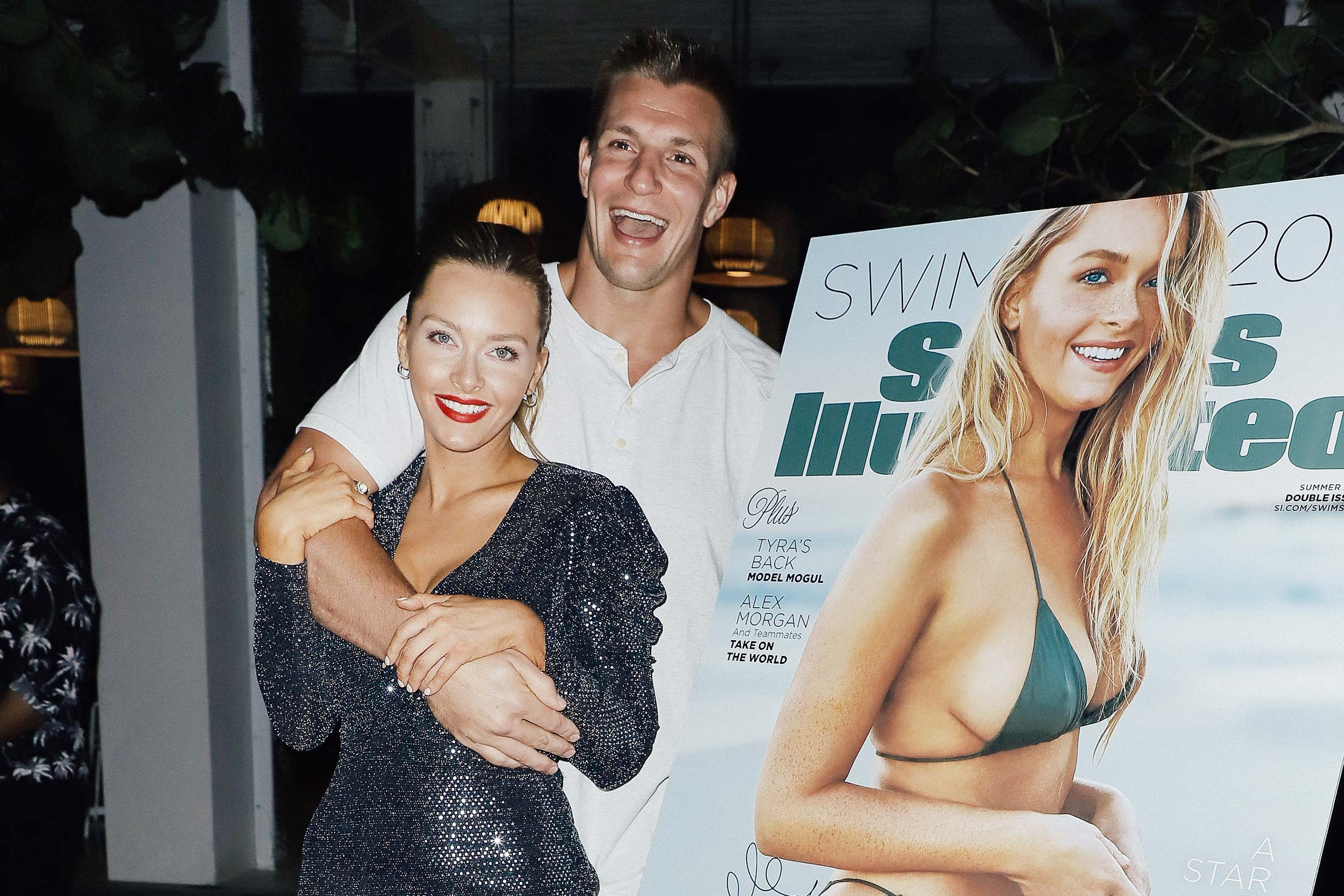 Rob Gronkowski and Camille Kostek embrace in front of a poster of her Sports Illustrated cover.