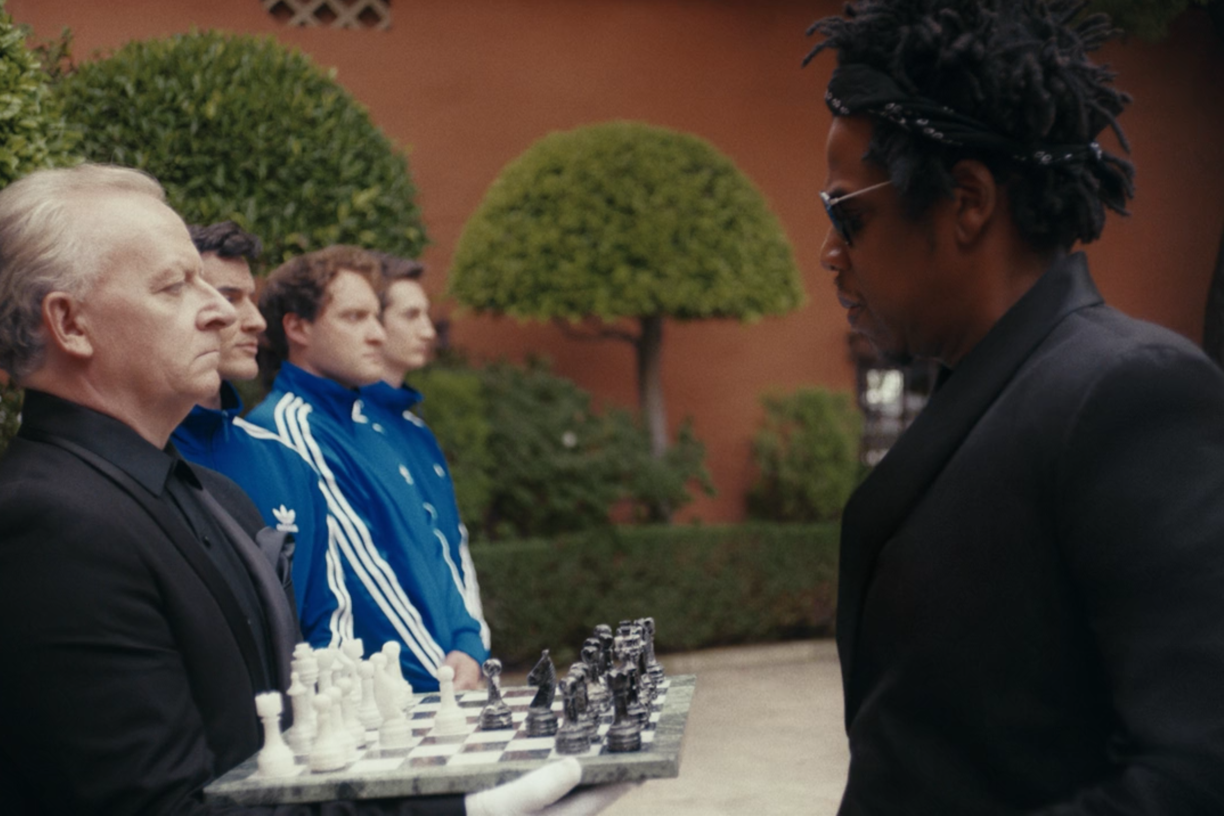 Jay-Z stands in front of a chessboard being held by a man in line with some other men.