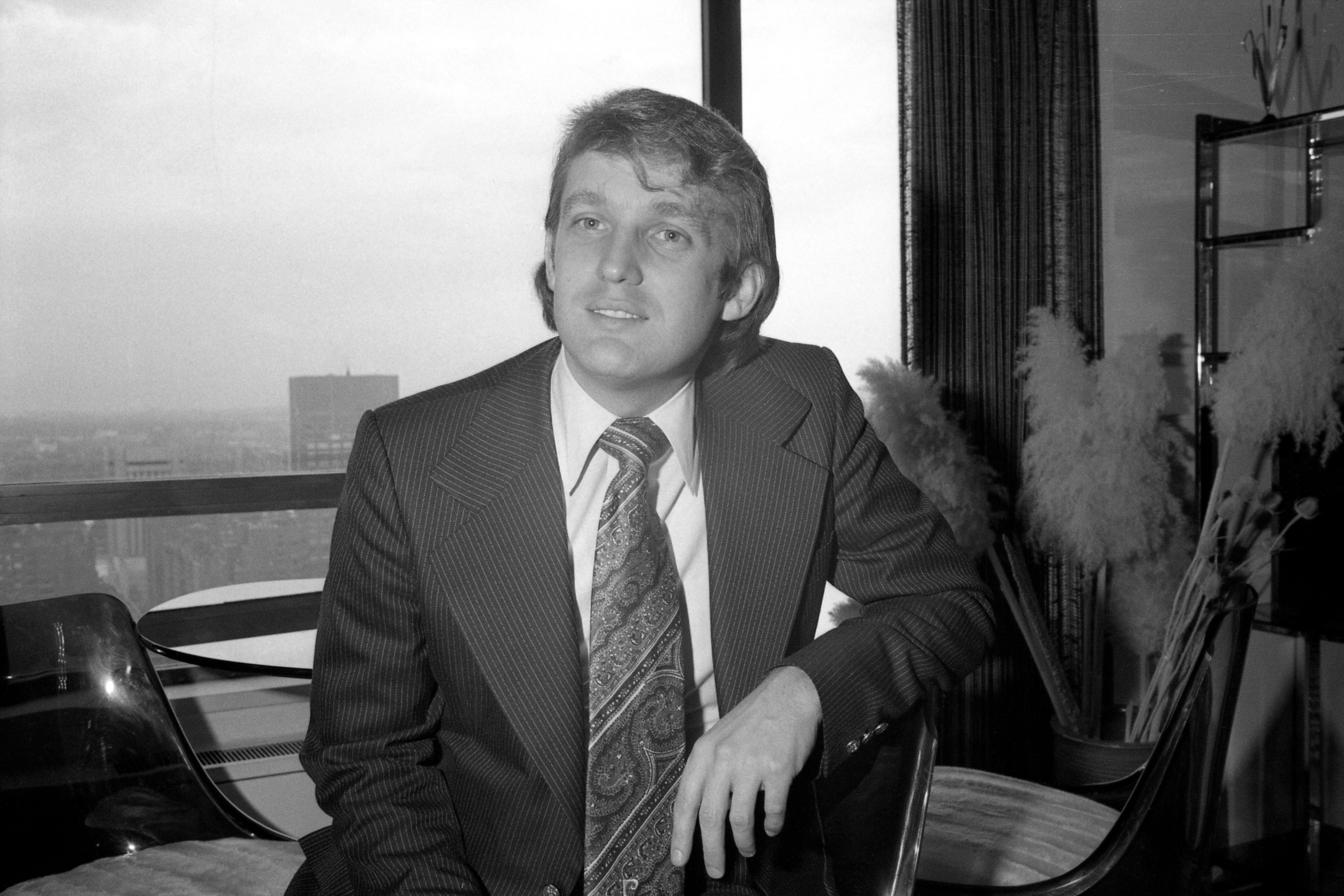 A young Trump wearing a suit and seated in front of a window in skyscraper.