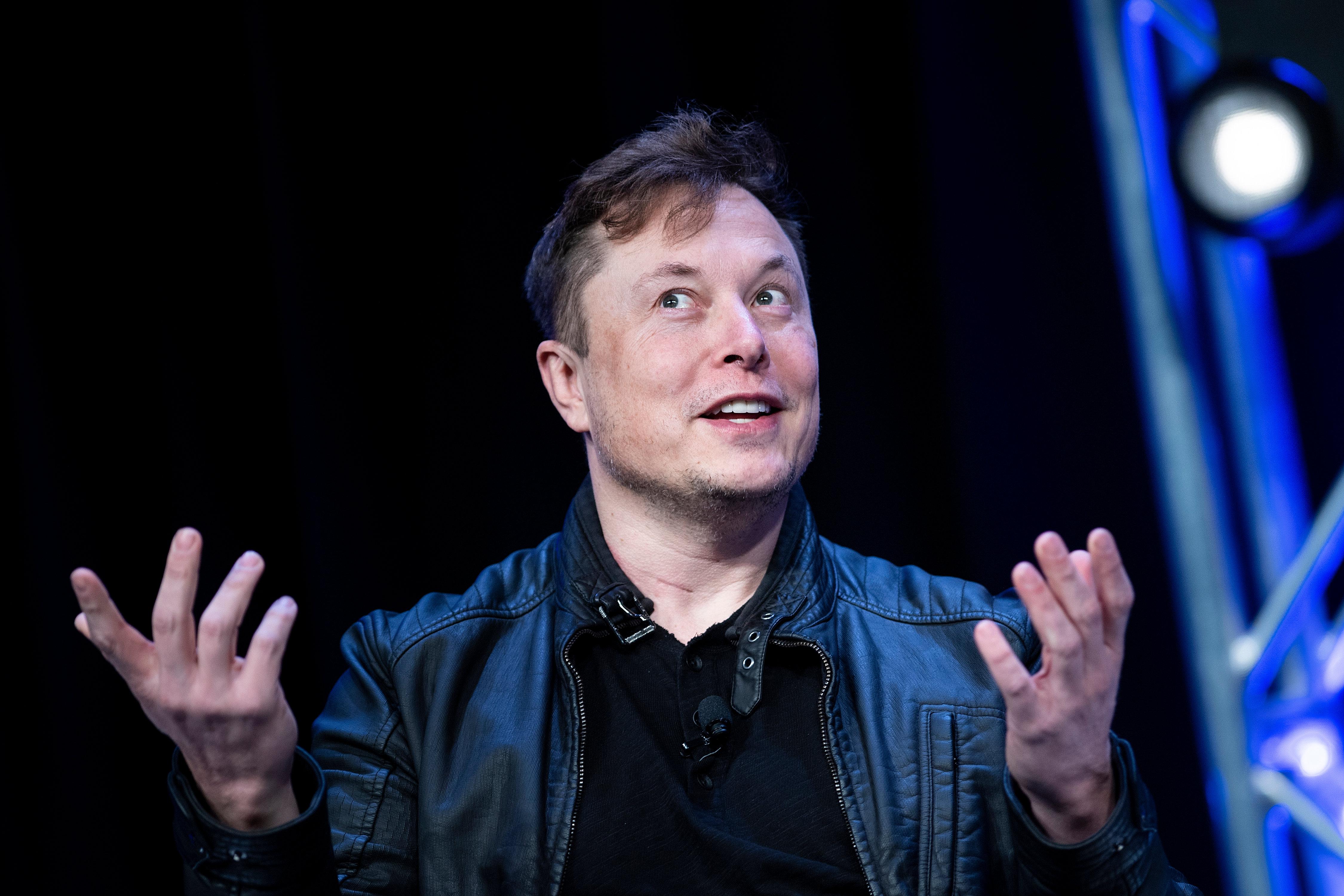 Elon Musk raises both hands and looks askance.