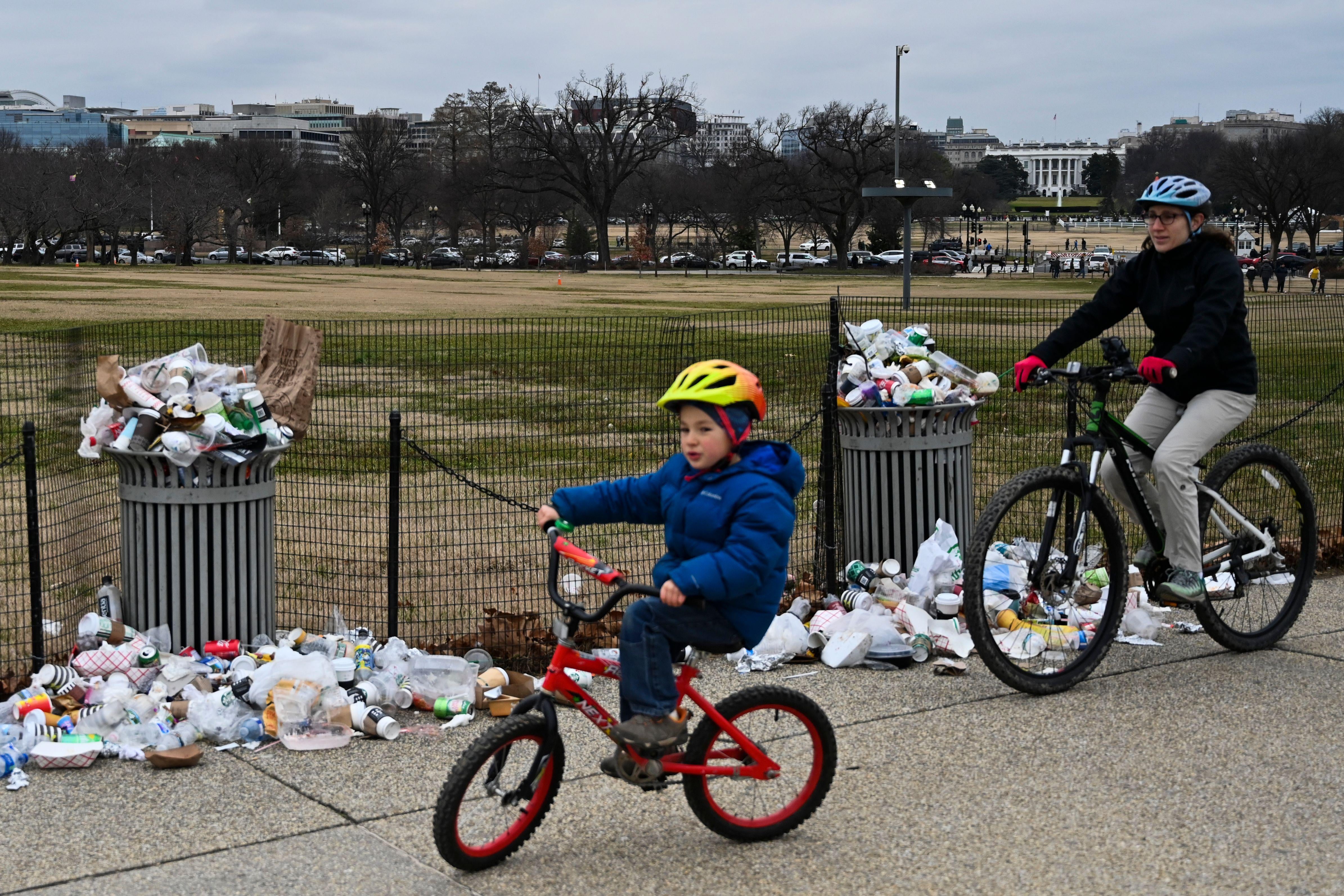 A child and adult on bicycles pass overflowing garbage cans. Behind them, the White House is distantly visible. 