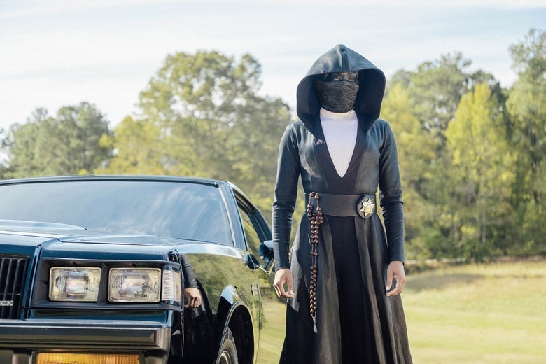 A black-cloaked figure wearing a mask stands next to a car.