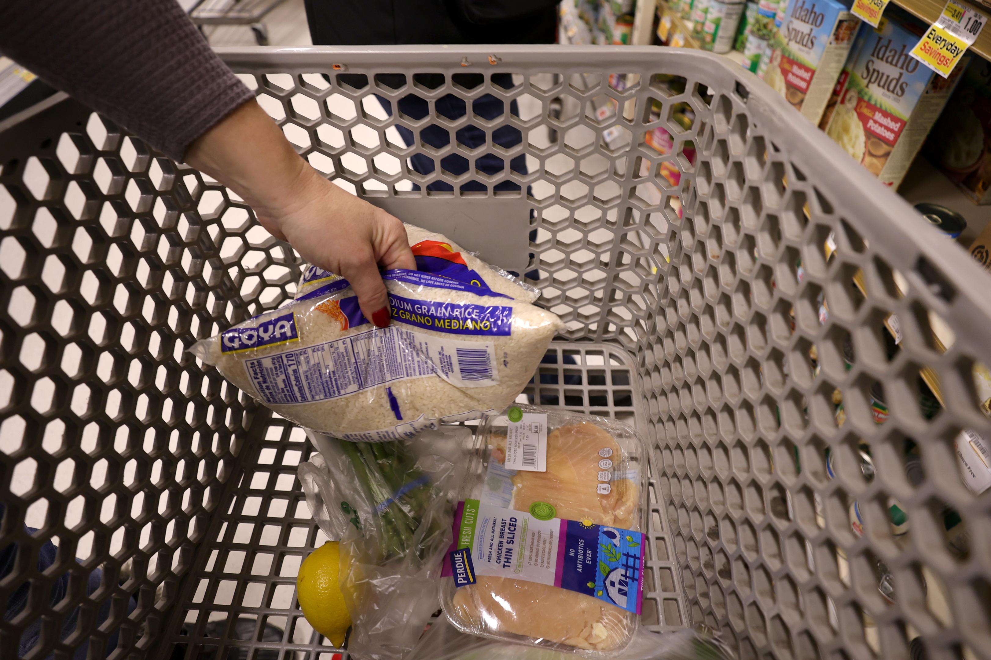 A hand putting a bag of rice into a grocery cart that already contains a lemon and a pack of chicken thighs