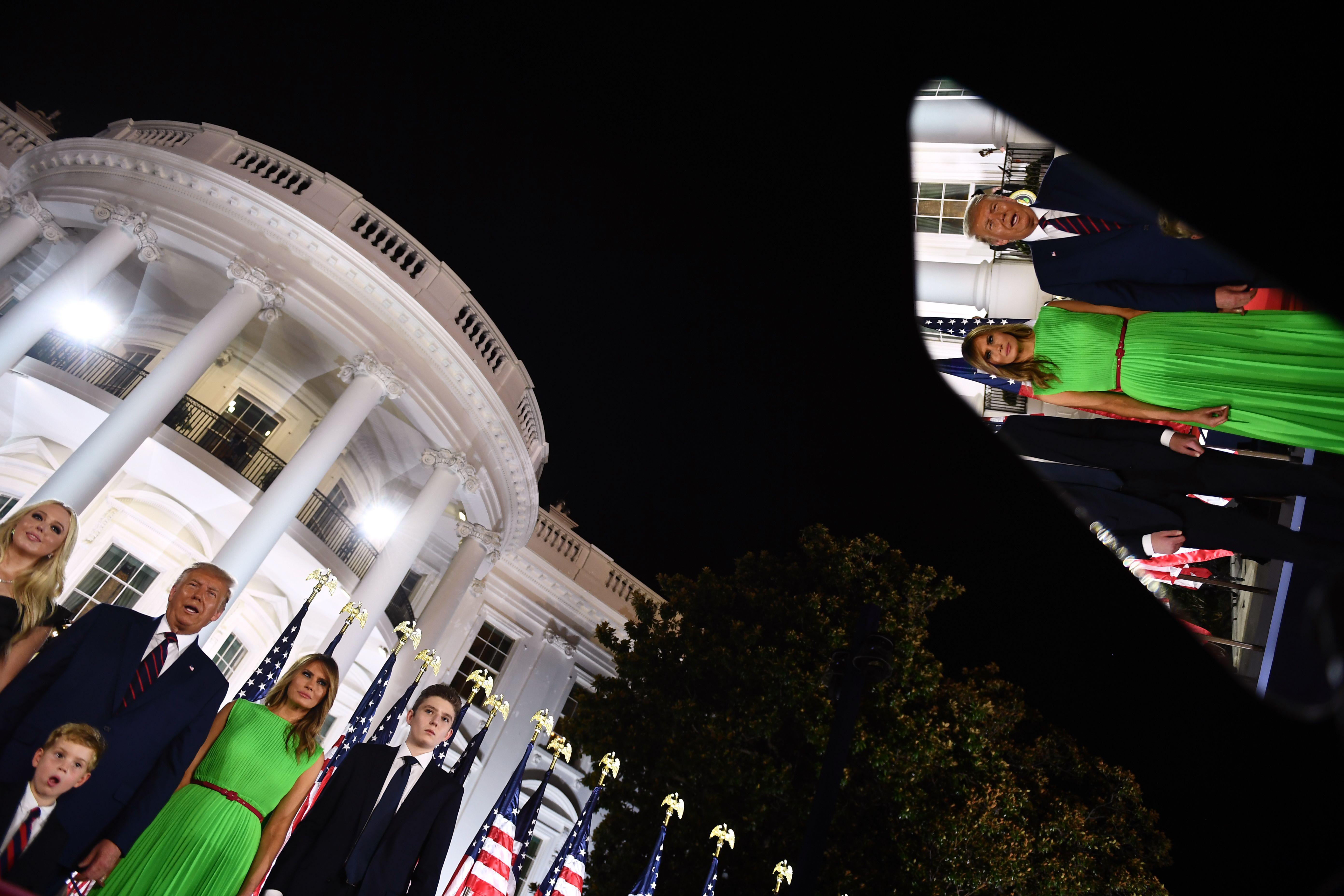 The Trump family stands in front of the White House at night. Their image is reflected in the screen of a teleprompter.