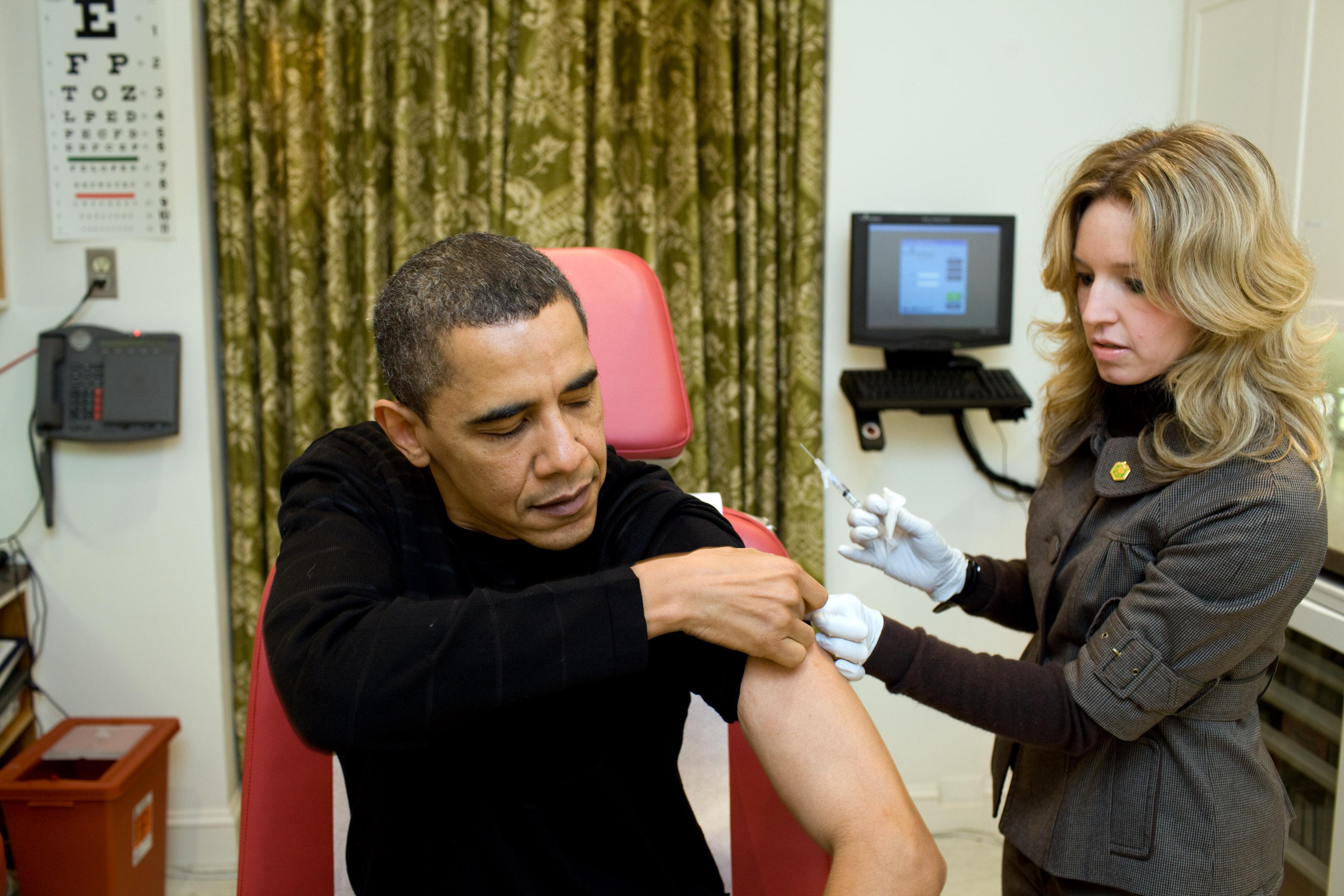 Barack Obama rolls up his sleeve as a woman holds a syringe.