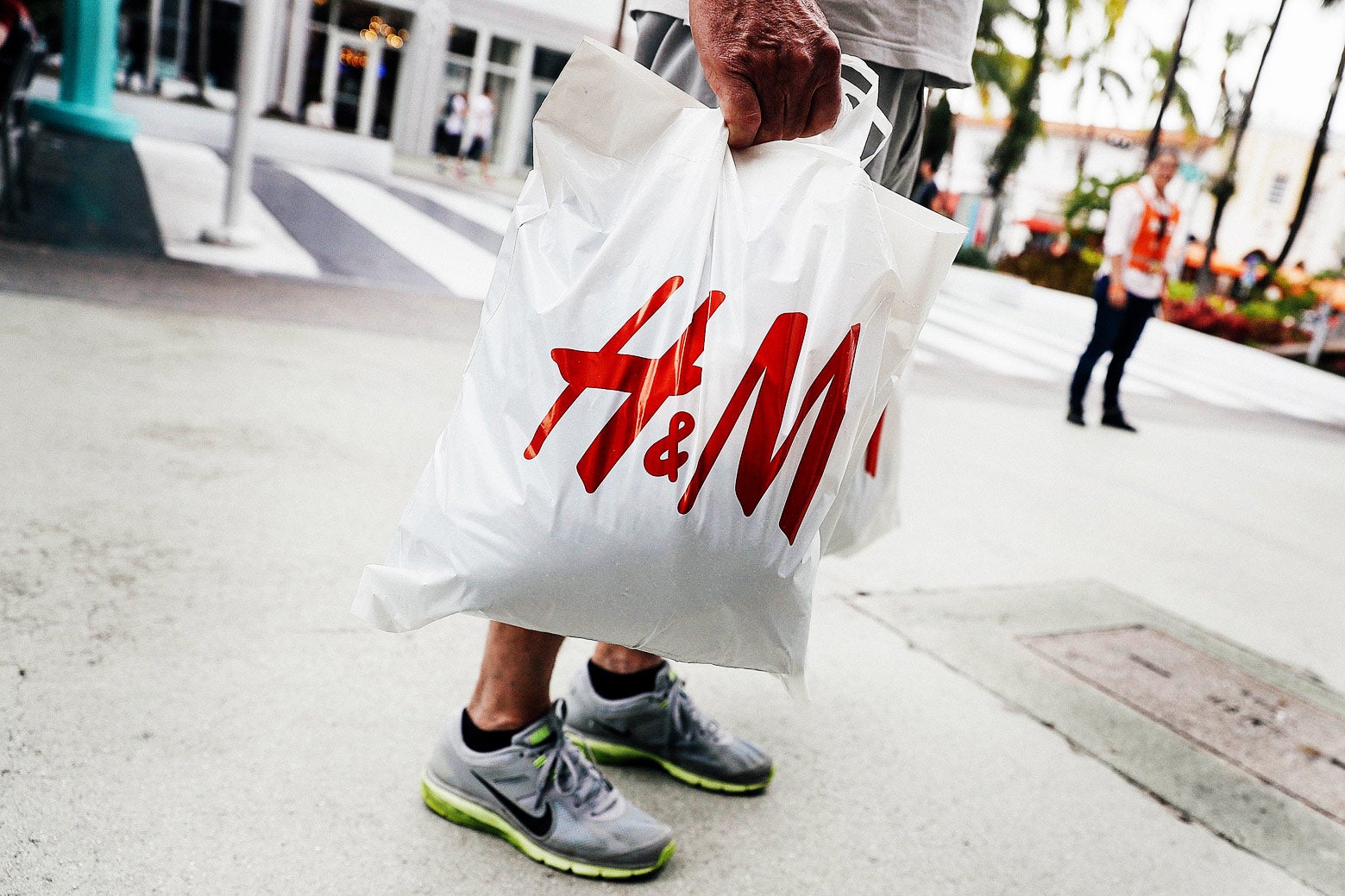 A plastic bag with the H&M logo on it, held by a man in shorts and sneakers outside a store in Miami.