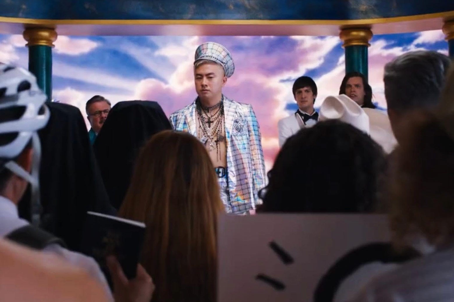 In the center, Bowen Yang wears a suit that looks like shiny graph paper, several gold necklaces, and a shiny-graph-paper hat, against a background of marble columns and pastel clouds. He appears to be addressing a crowd, and behind him on each side, the other actors look on.