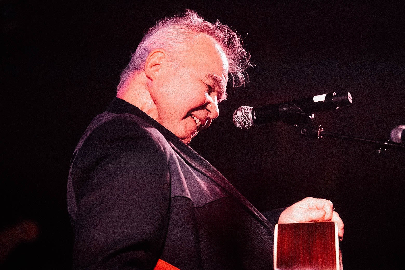 John Prine smiling as he performs with his guitar onstage