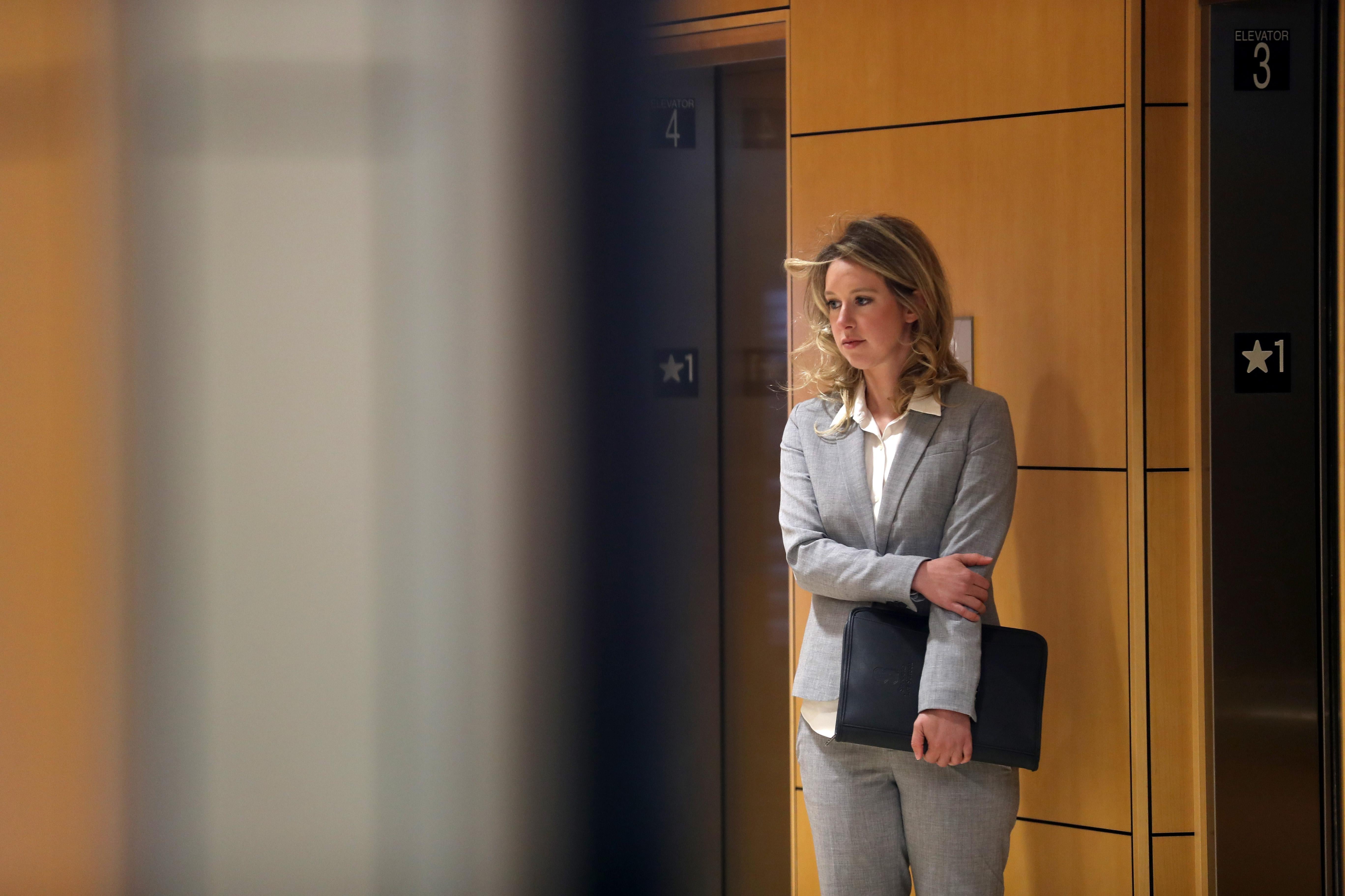 A woman in a gray suit stands in front of elevators, holding a portfolio, with one arm crossed in front of her.