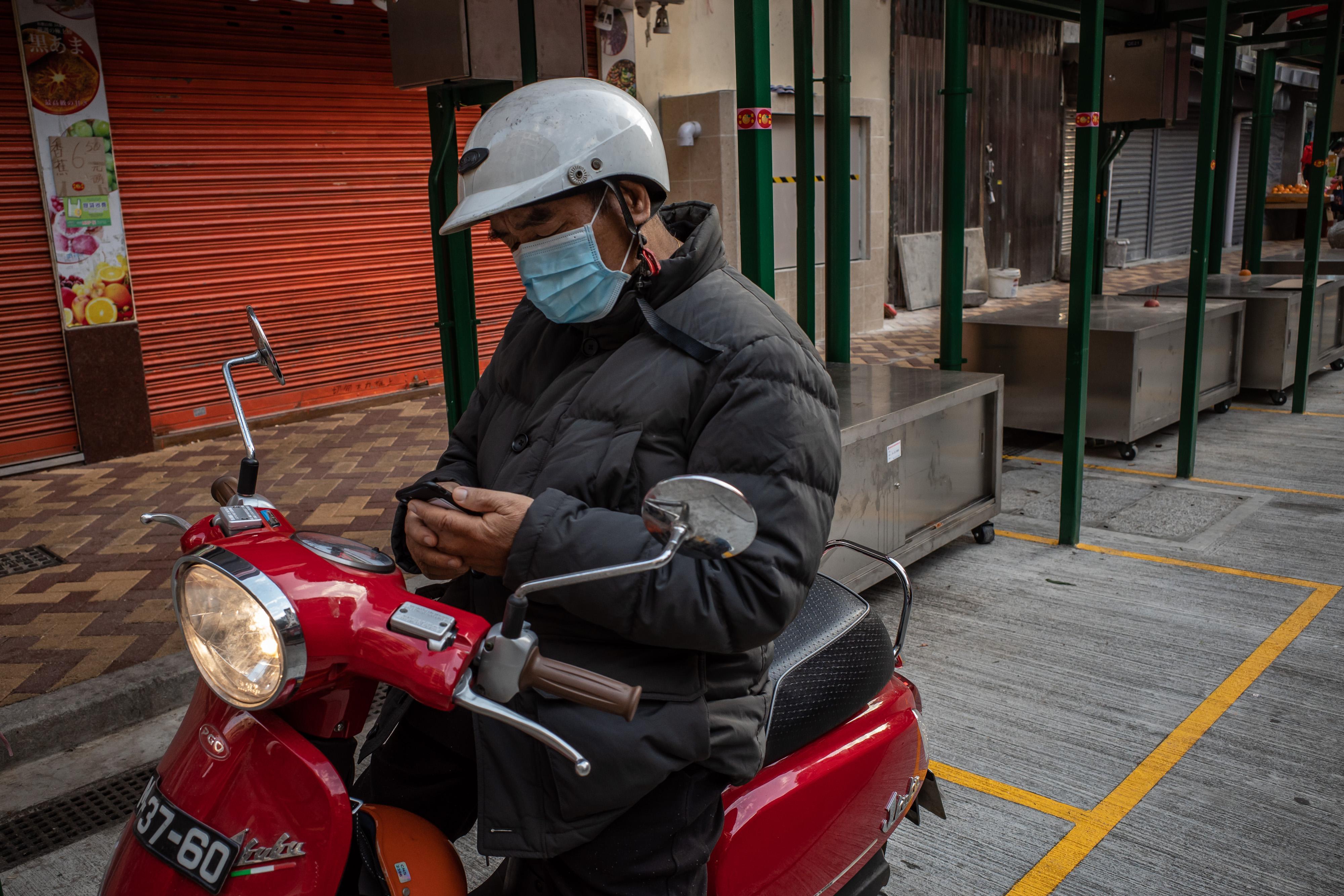 A motorcyclist wearing a face mask looks at his phone while sitting on his bike.