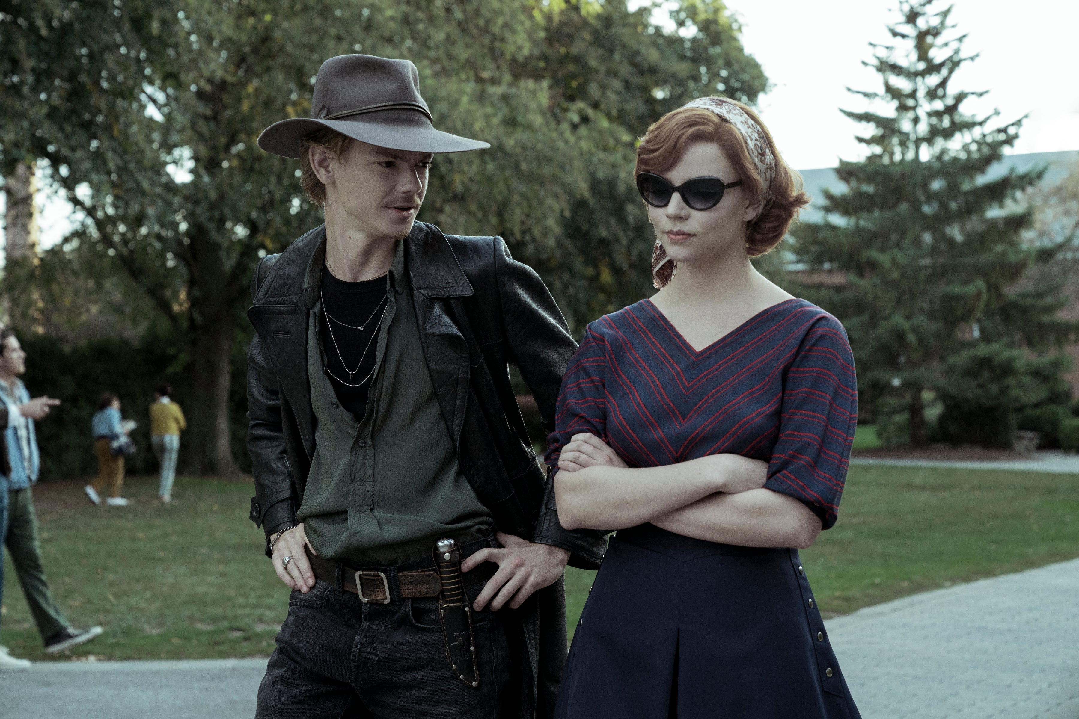 A man stands, hands on hips, in a cowboy hat and leather trenchcoat, next to a skeptical woman in sunglasses.