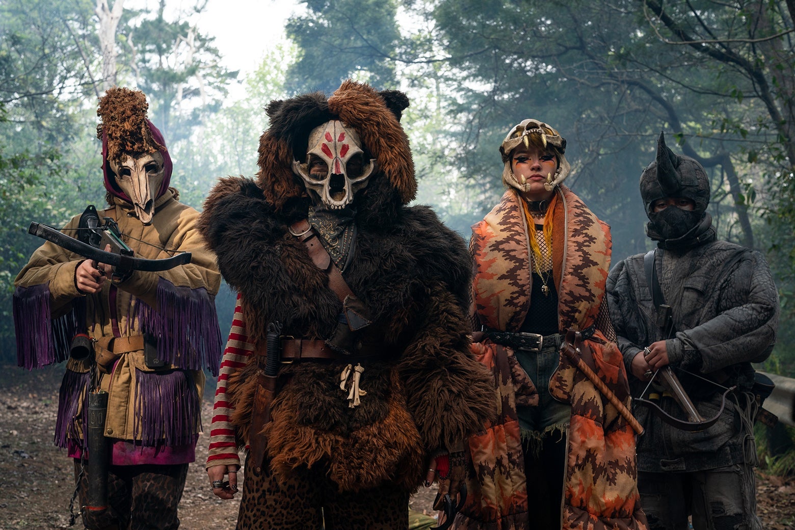 Four children, dressed in fur and wearing animal skulls over their faces, face the camera. The leftmost is aiming a crossbow at the camera.
