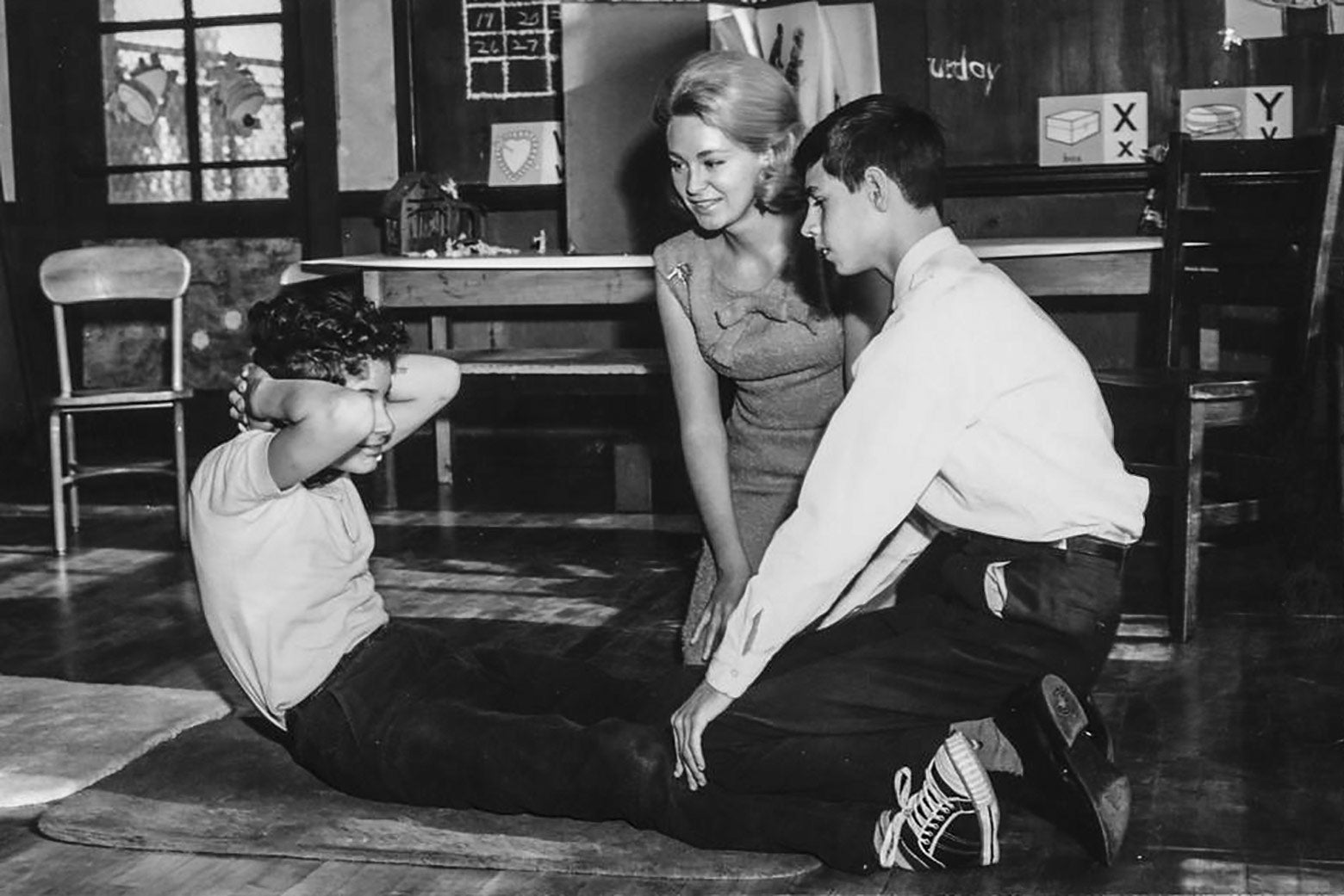 A black-and-white photo of a woman kneeling behind a duo of kids, one performing a situp and the other holding his legs steady, in a classroom.