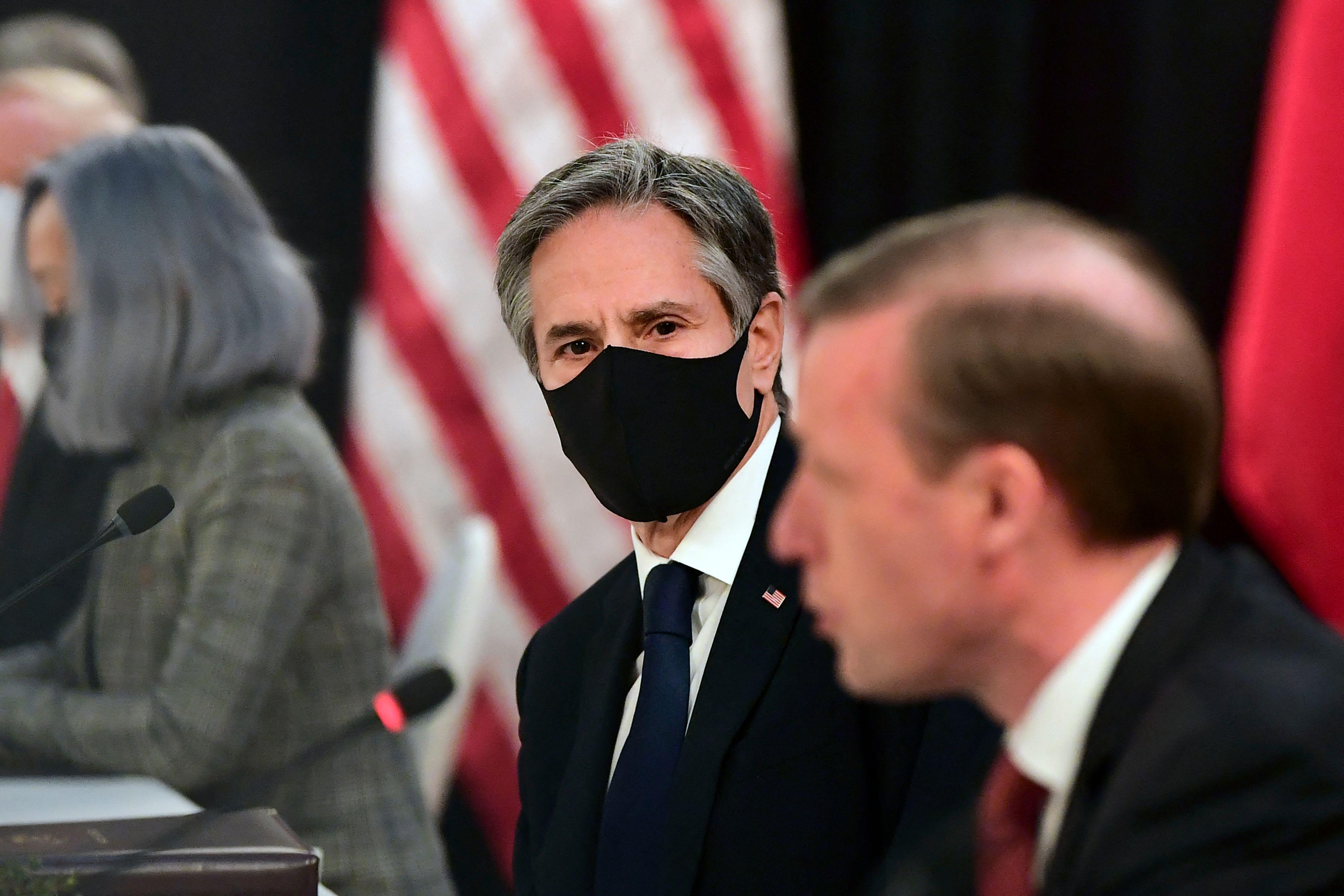 One man in the foreground speaking. Another in the background, in a mask, looking on.