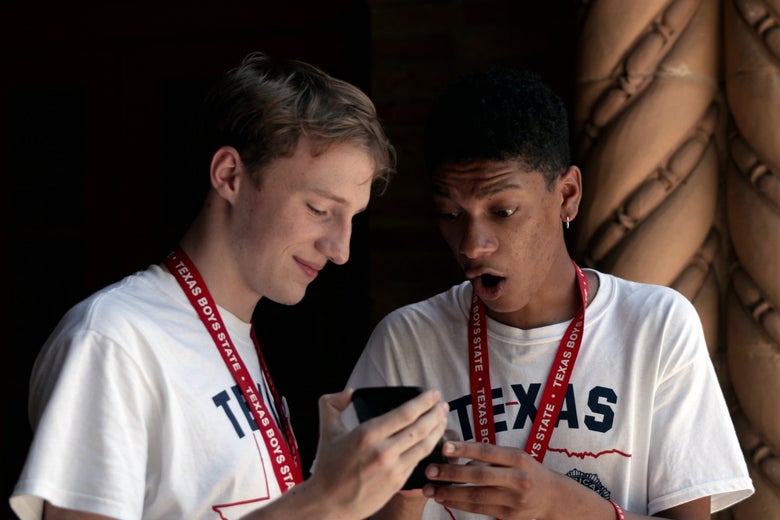 A white boy holds out his cellphone to a Black boy, who looks at the screen, mouth agape. Both are wearing Texas Boys State shirts and lanyards.