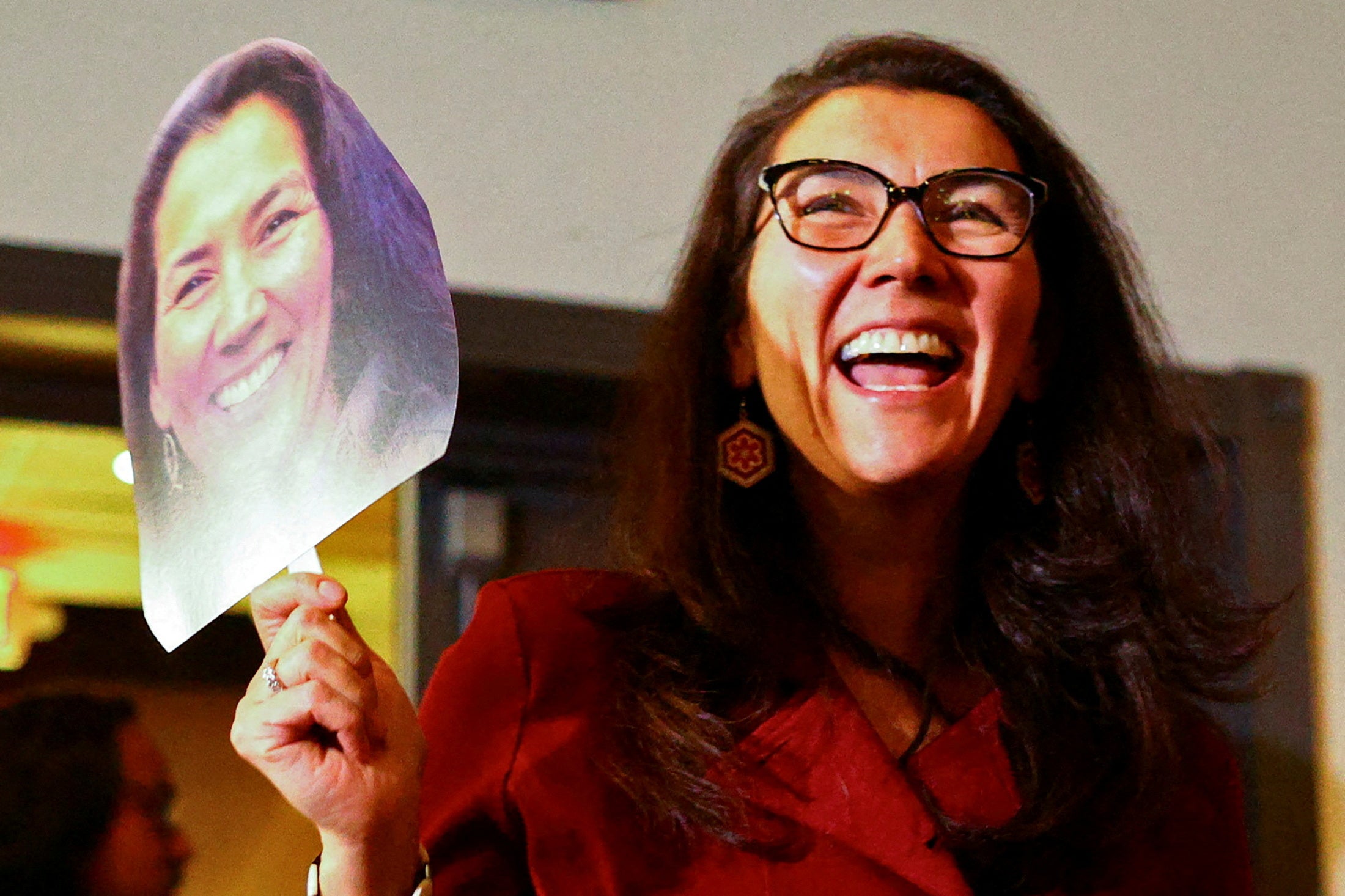 A woman smiling widely, holding a mask decorated with an image of her face.